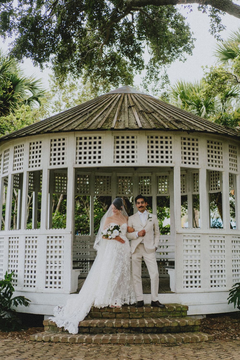 White latticed wedding gazebo in a garden setting surrounded by lush trees and greenery.