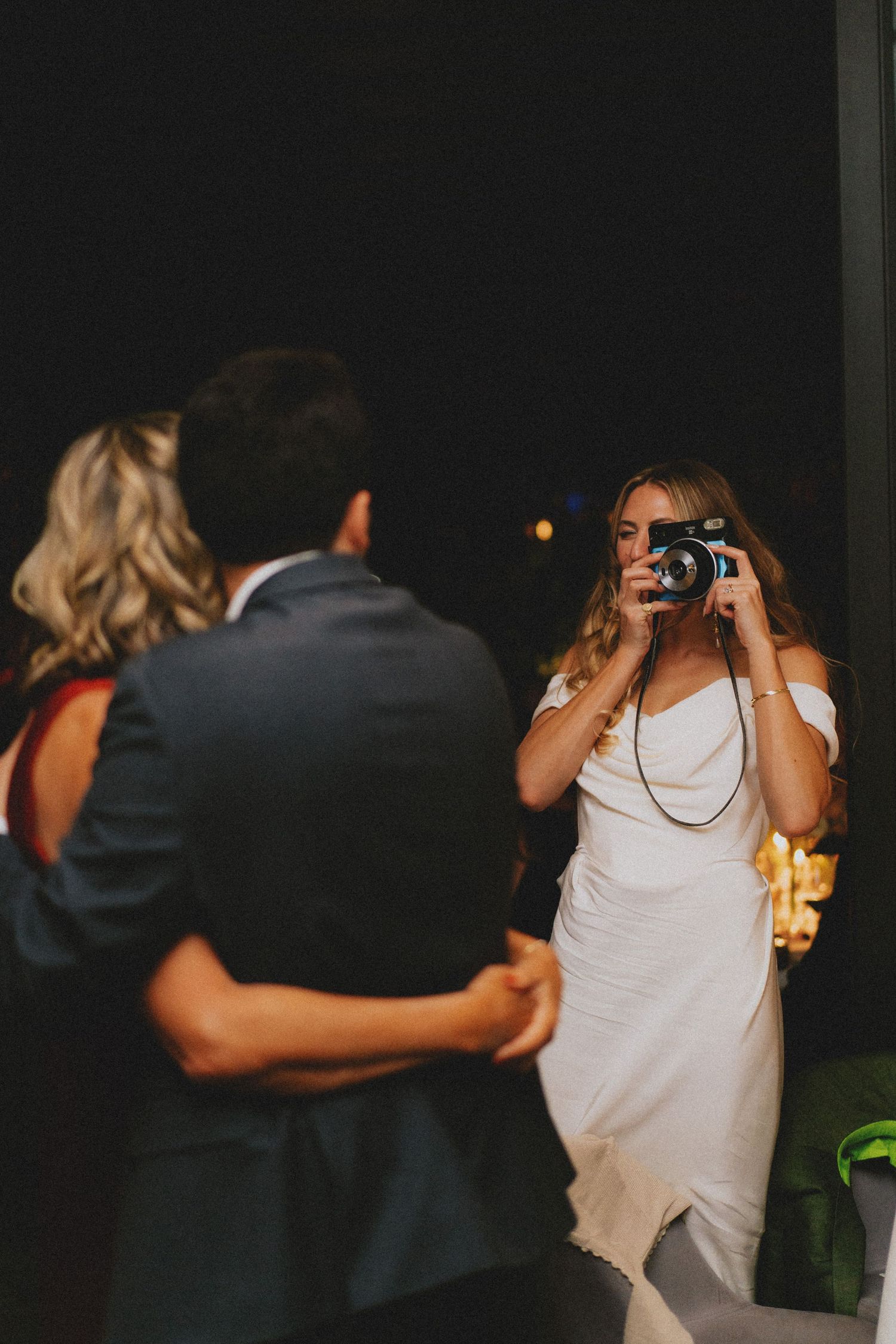 Bride in white gown photographs dancing couple with film camera during wedding reception.