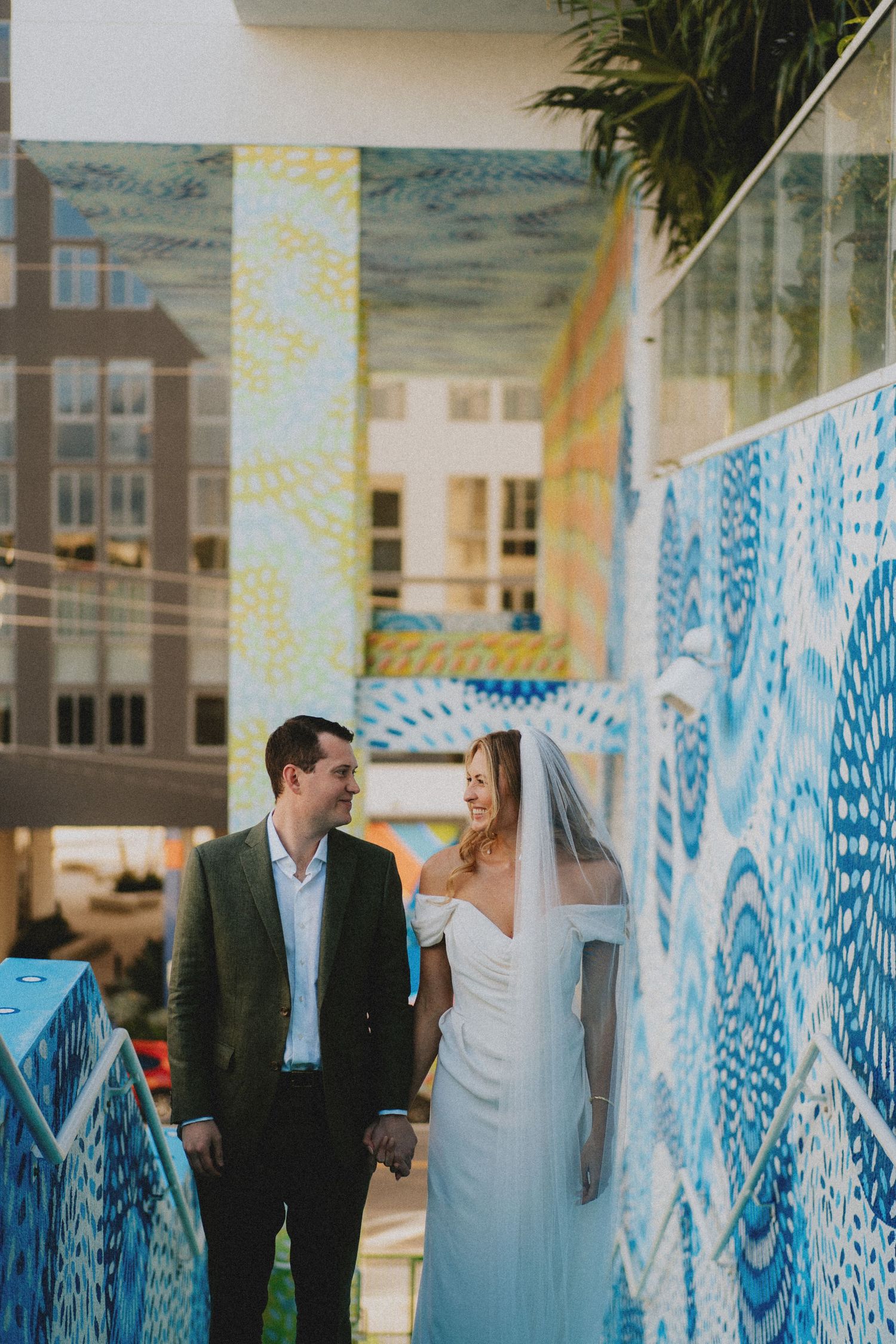 Bride and groom hold hands beside a colorful blue mosaic wall at their outdoor wedding venue.