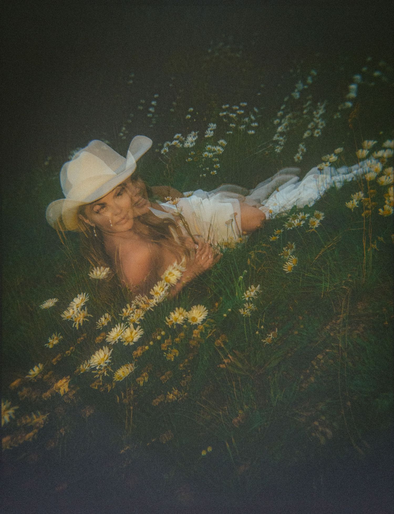 Woman in white cowboy hat lying among wildflowers in a field, wearing white outfit, dreamy film photography aesthetic.