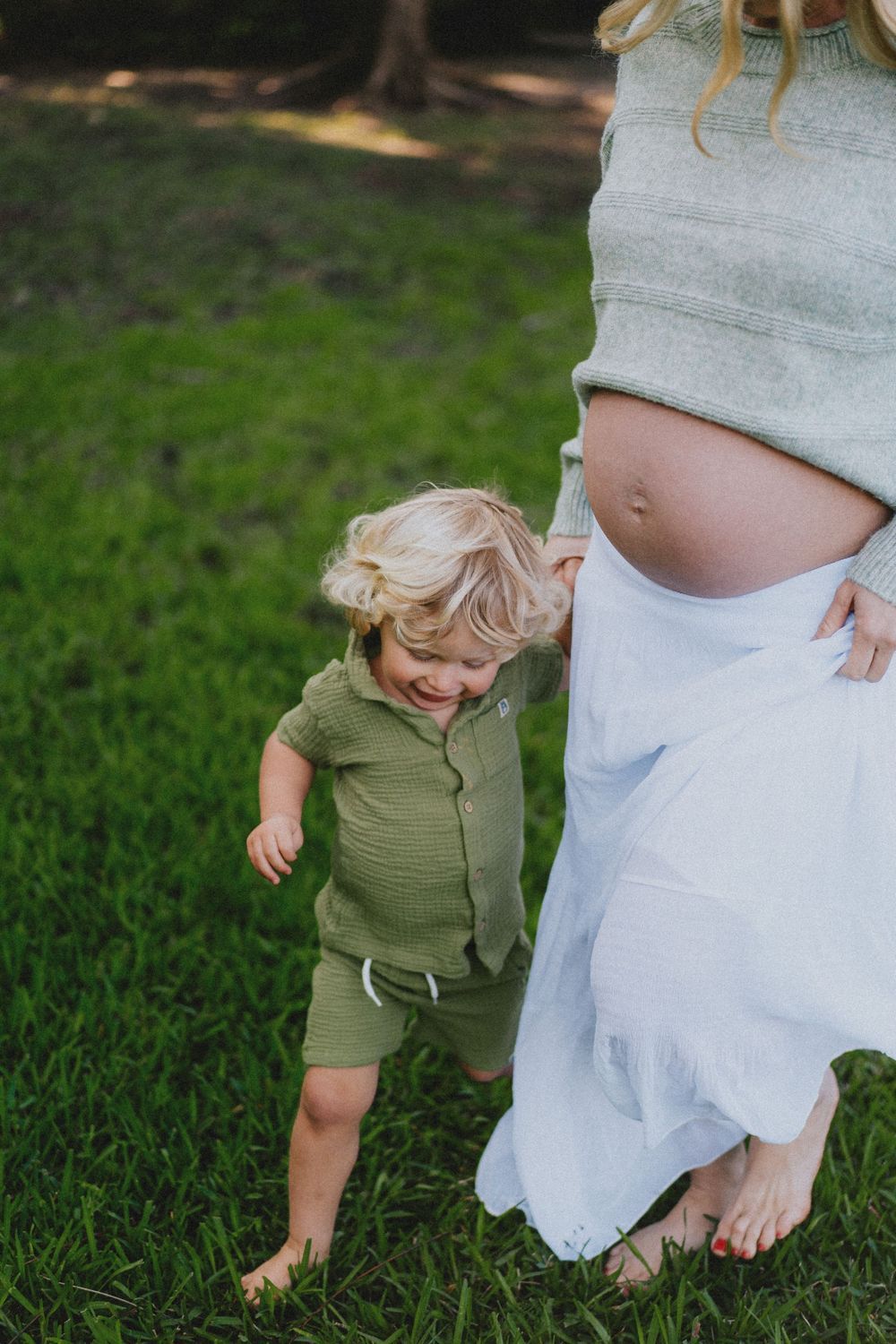 A child in olive green plays in the grass next to someone wearing a flowing white dress.