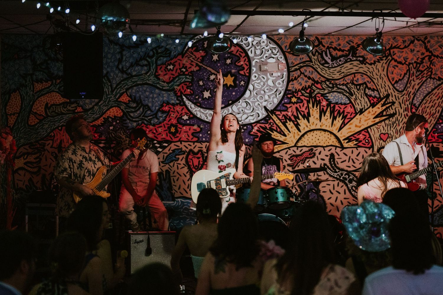 Band performing live on stage with colorful psychedelic mural backdrop, lead singer raising arm, crowd watching.