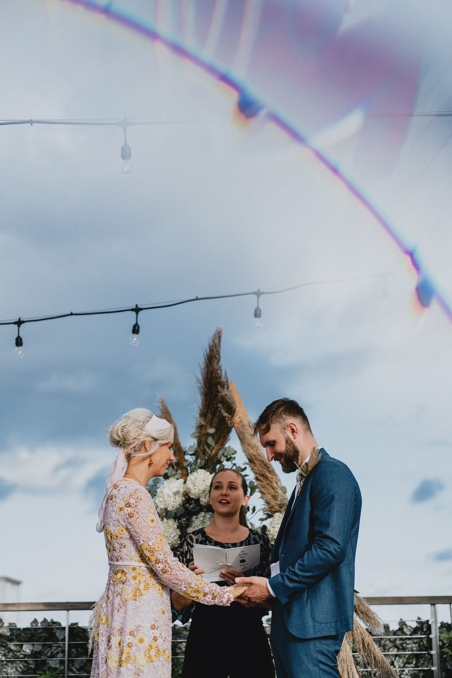 Outdoor wedding ceremony with bride in floral dress and groom in blue suit exchanging vows under string lights.