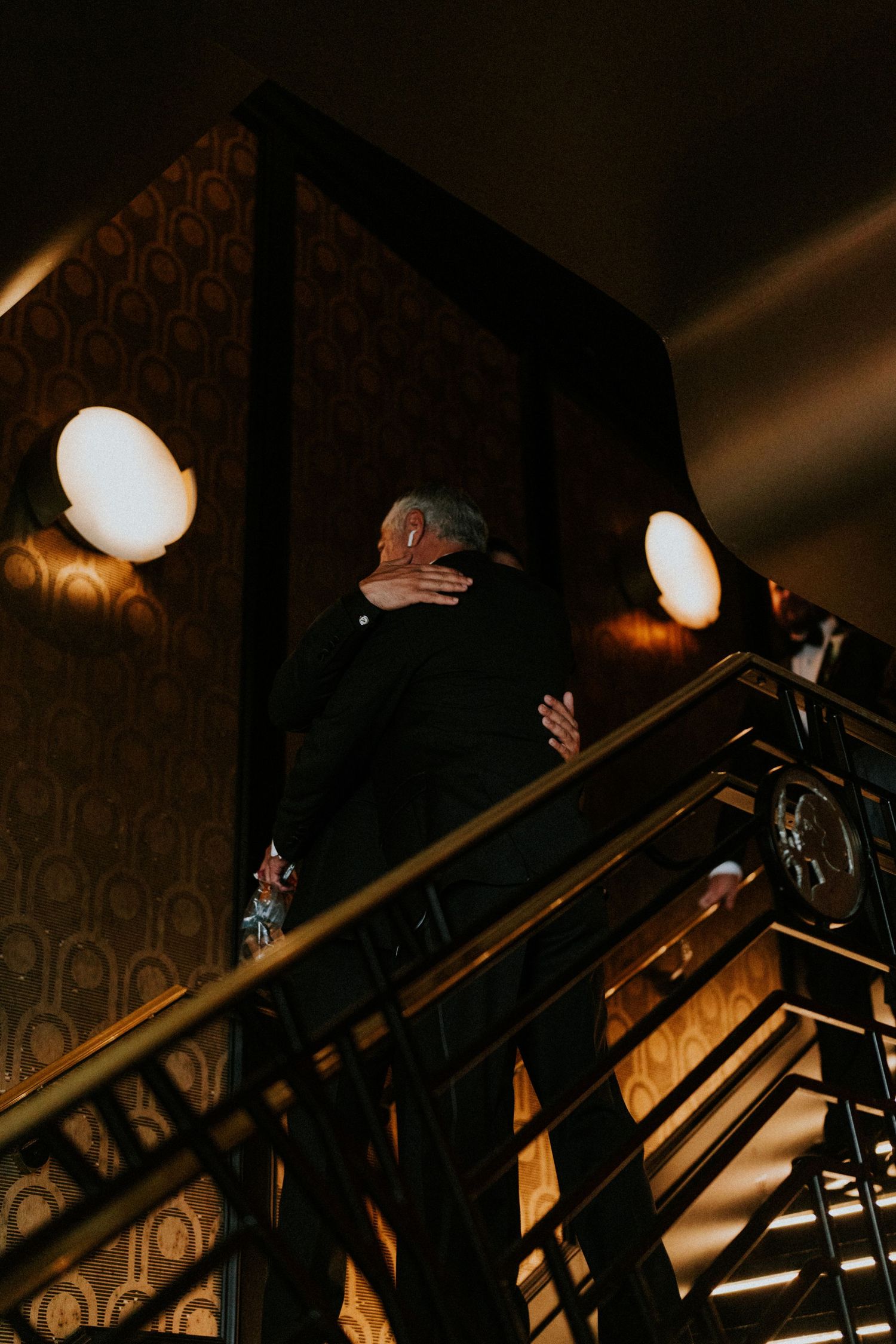 Two people embrace at the top of an elegant staircase, lit by warm wall sconces in a moody interior.
