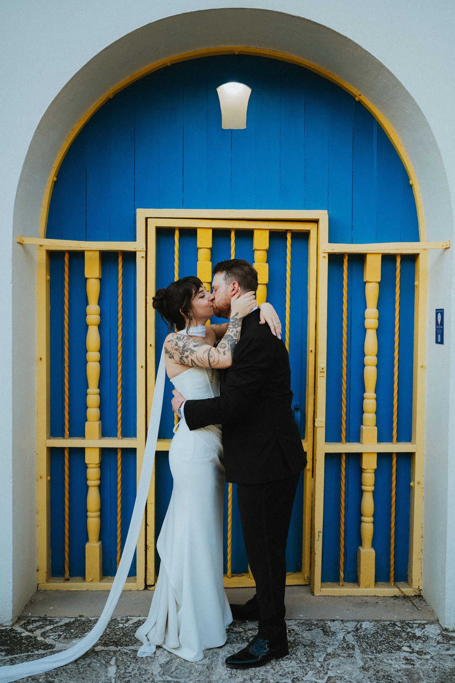 Bride and groom share a romantic kiss in front of a blue and yellow arched doorway.