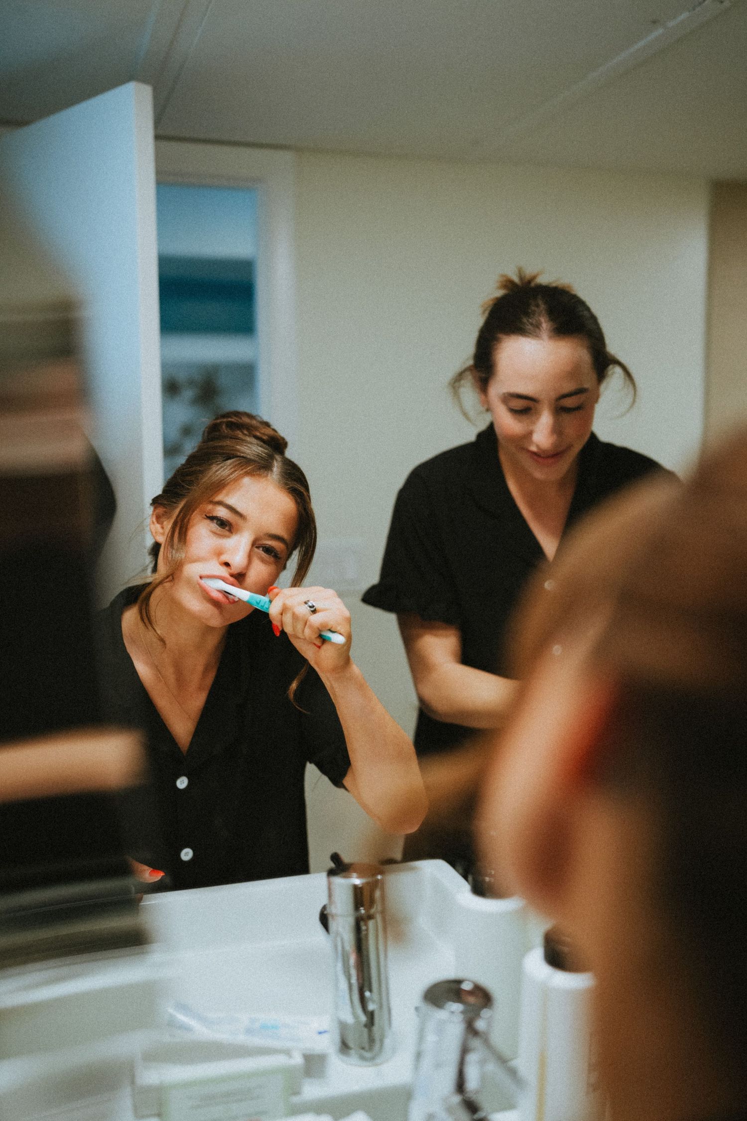 Two women in black pajamas brushing teeth together in a bright bathroom mirror reflection.