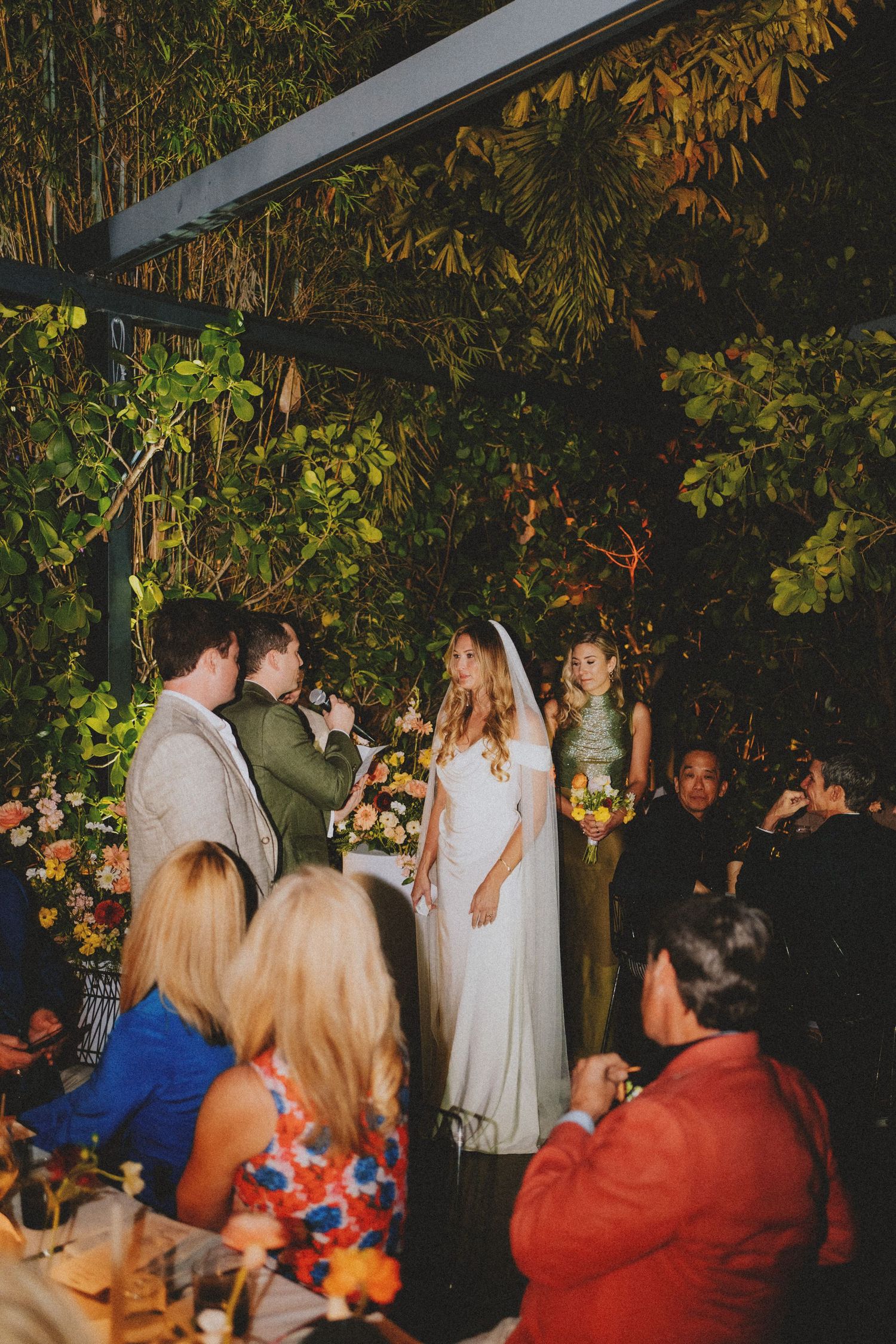 Bride and groom exchange vows at outdoor garden wedding ceremony surrounded by lush greenery and floral arrangements.