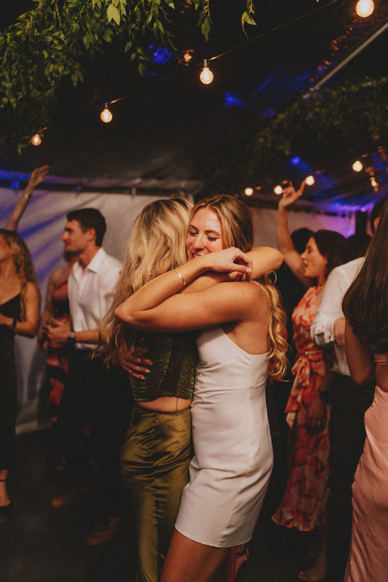 Two blonde women in elegant dresses share a warm hug on the dance floor at a wedding reception.