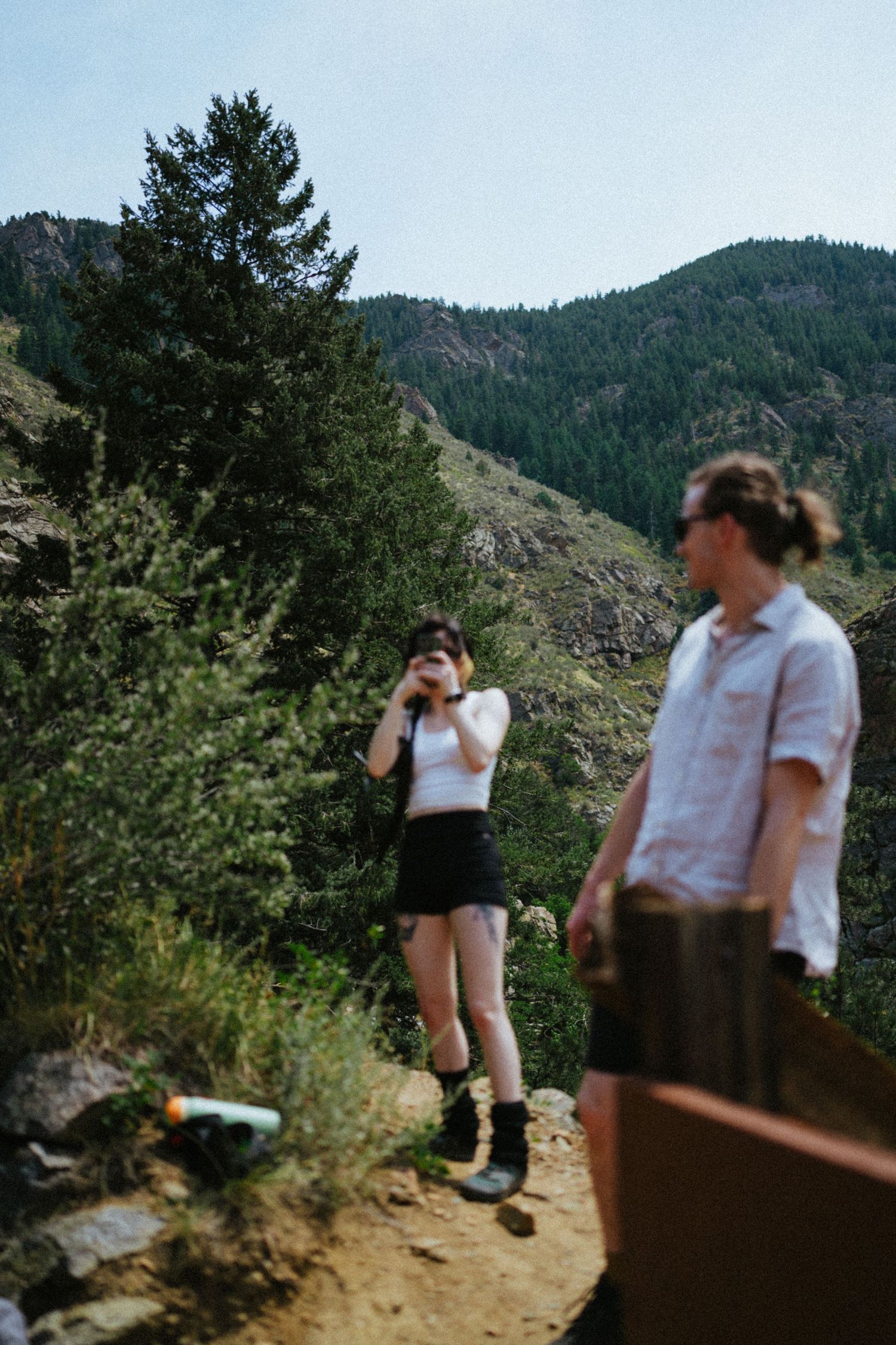 Two hikers on a mountain trail, one photographing scenic forested peaks while another stands nearby in casual outdoor attire.