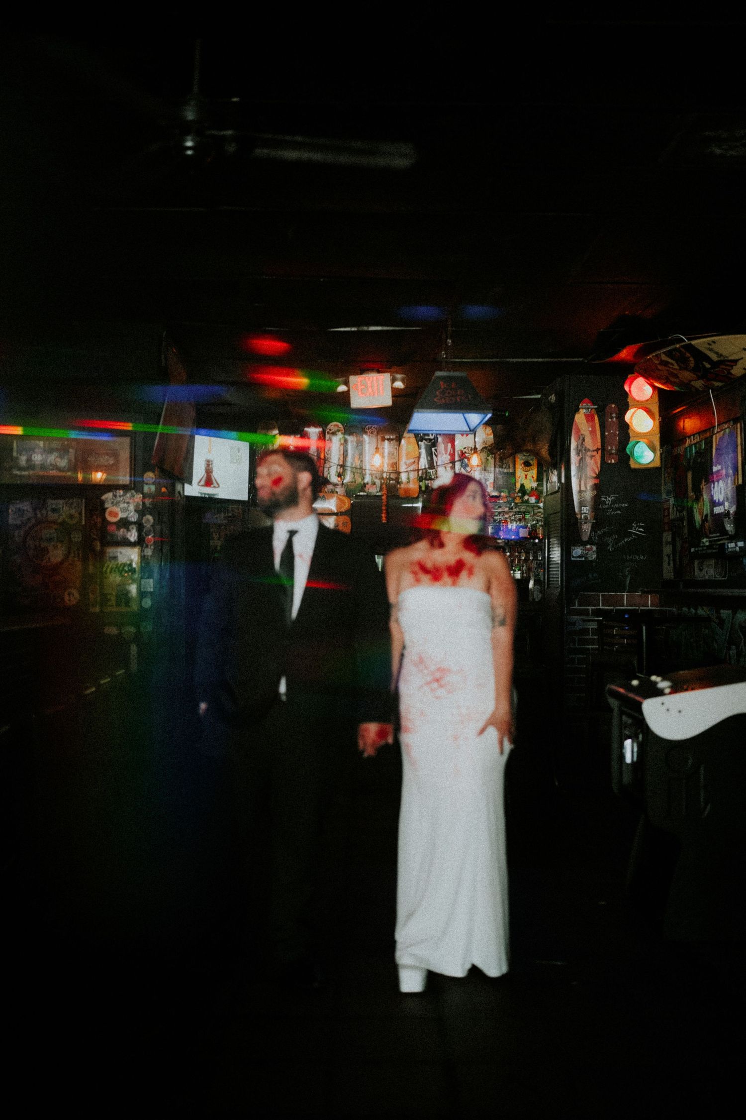 Zombie wedding couple holding hands in dark neon-lit bar, bride in blood-stained white dress, groom in black suit.