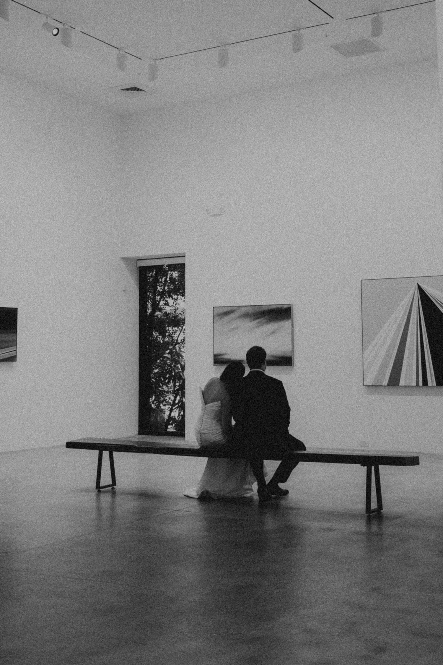 Black and white photo of a couple in wedding attire sitting on a bench in an art gallery.