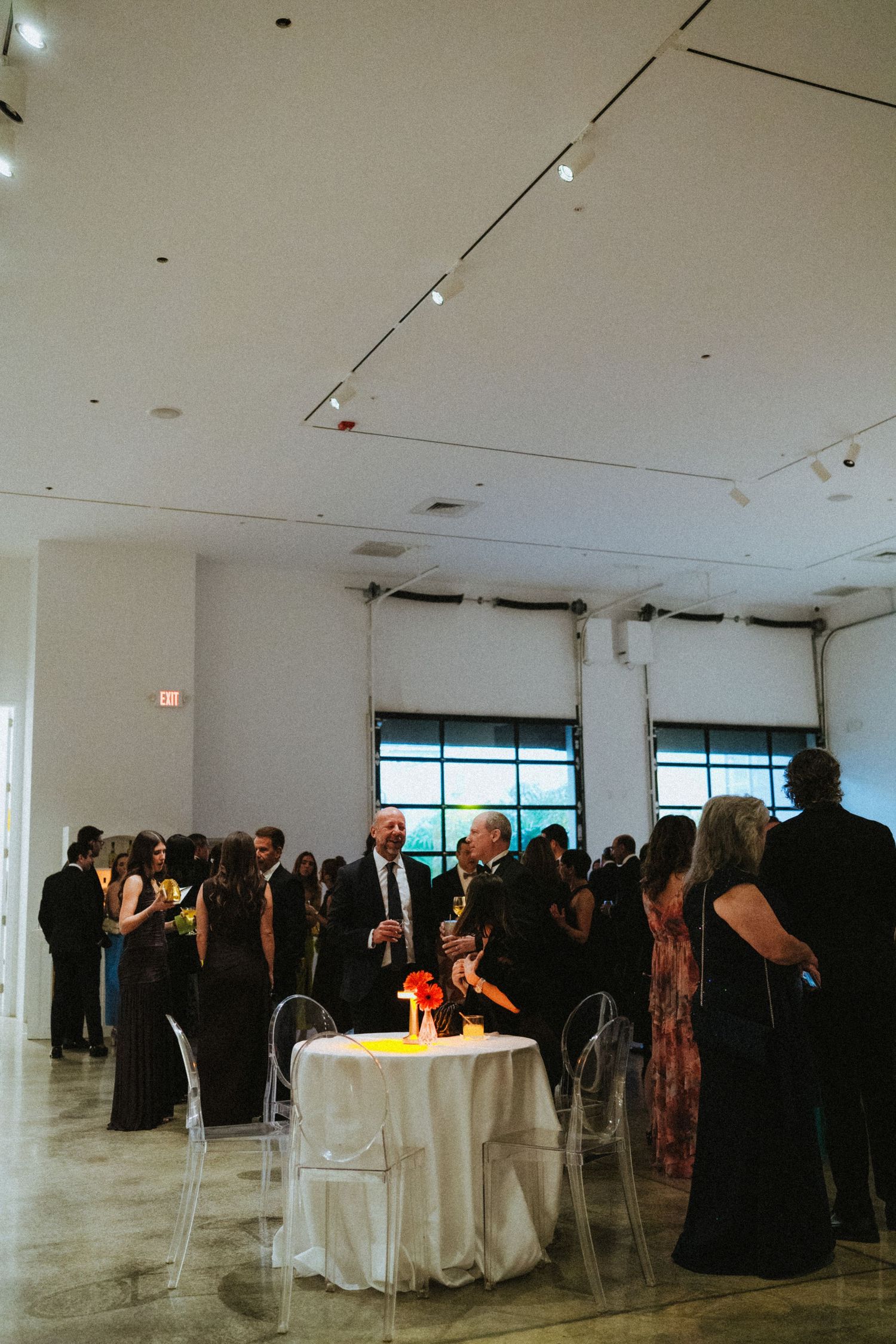Elegant cocktail hour in a modern white gallery venue with guests mingling around ghost chairs and candlelit tables.