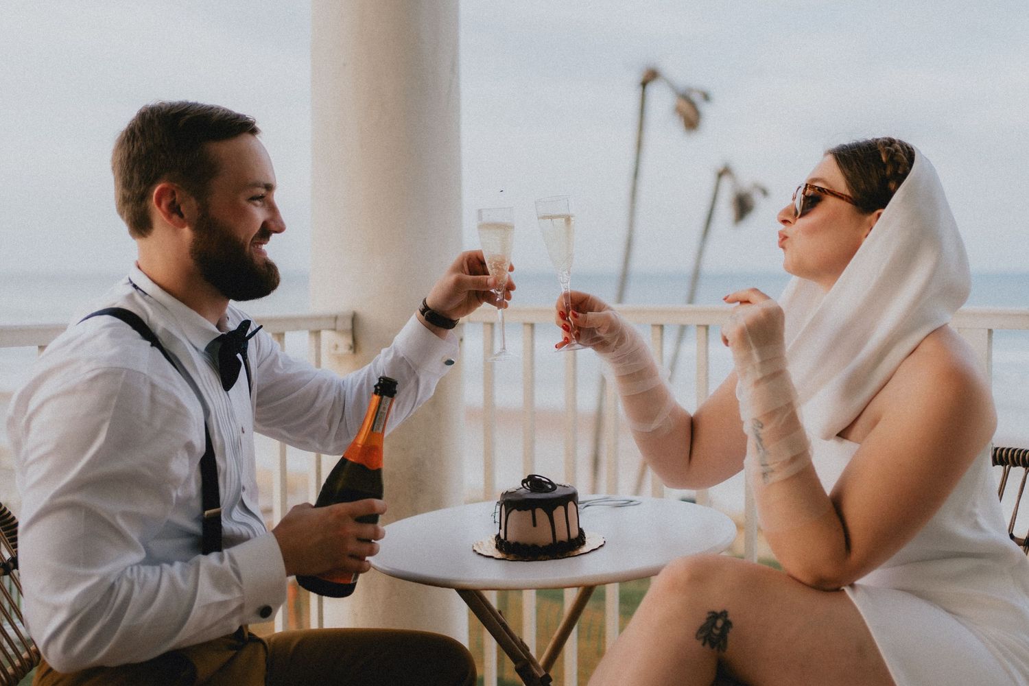 Bride and groom toasting champagne at a seaside balcony table with a small wedding cake.