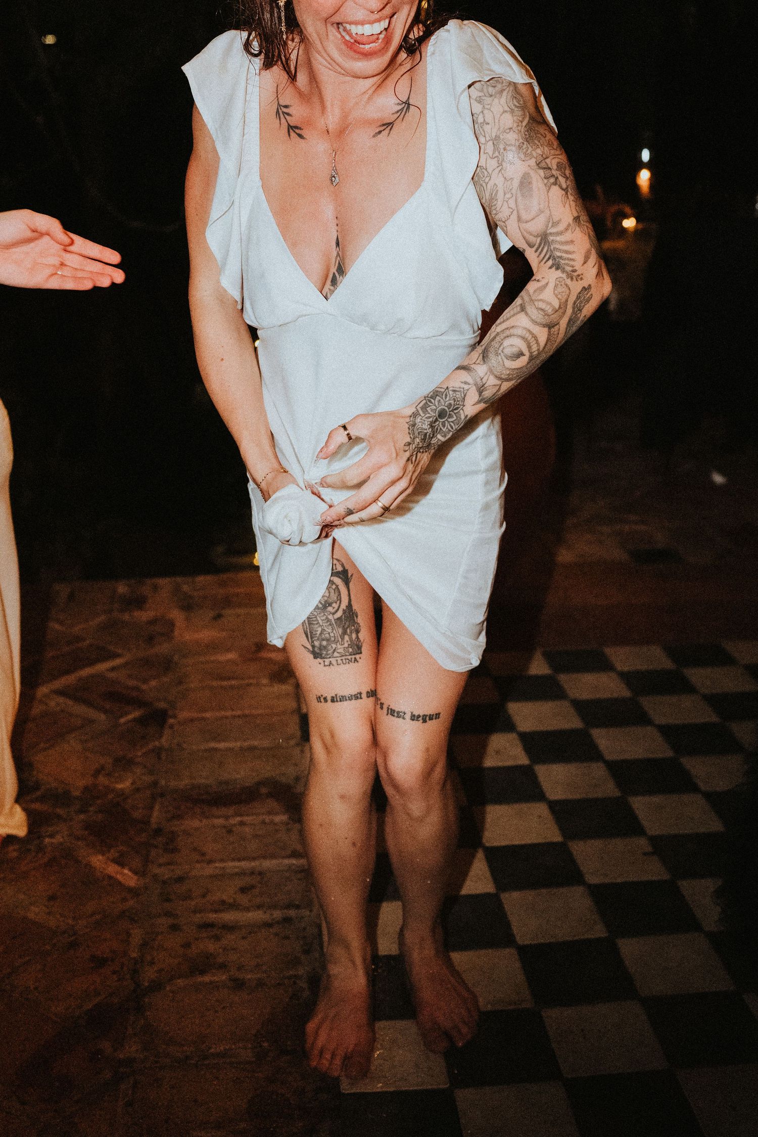 Tattooed bride in white dress dancing barefoot on checkered floor at outdoor wedding reception at night.