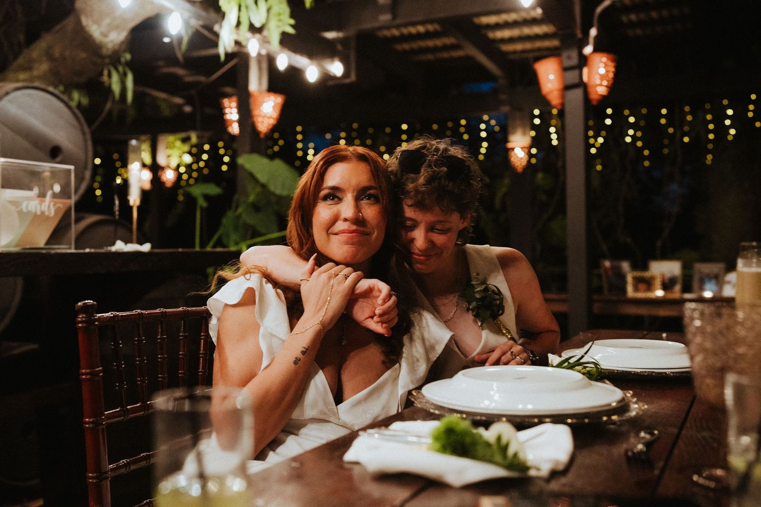 Two brides share a tender embrace at a candlelit wedding reception table, surrounded by warm string lights.