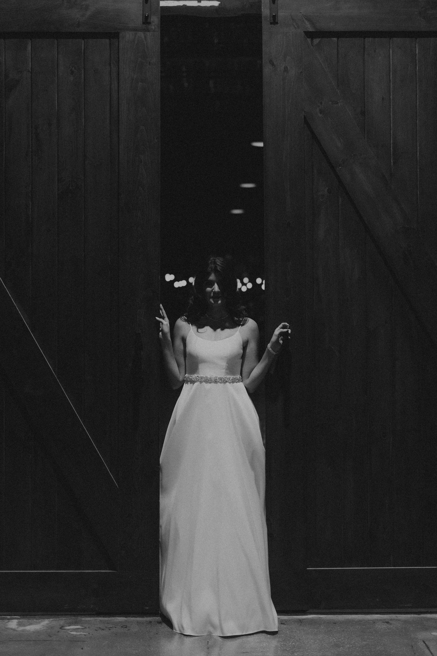 Black and white photo of a bride in a white gown standing between rustic barn doors with floral hair accessories.