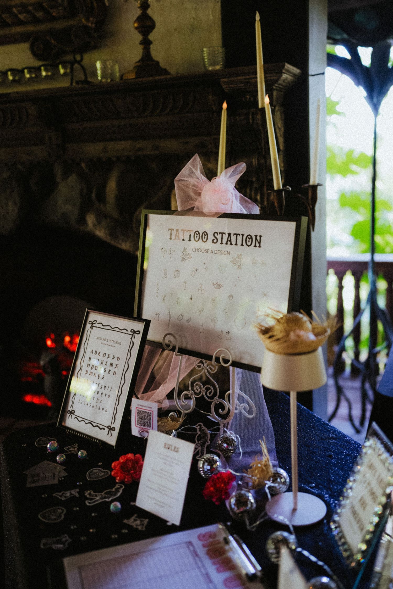 Elegant tattoo station display at a wedding reception with candles, framed sign, and decorative accessories near a fireplace.