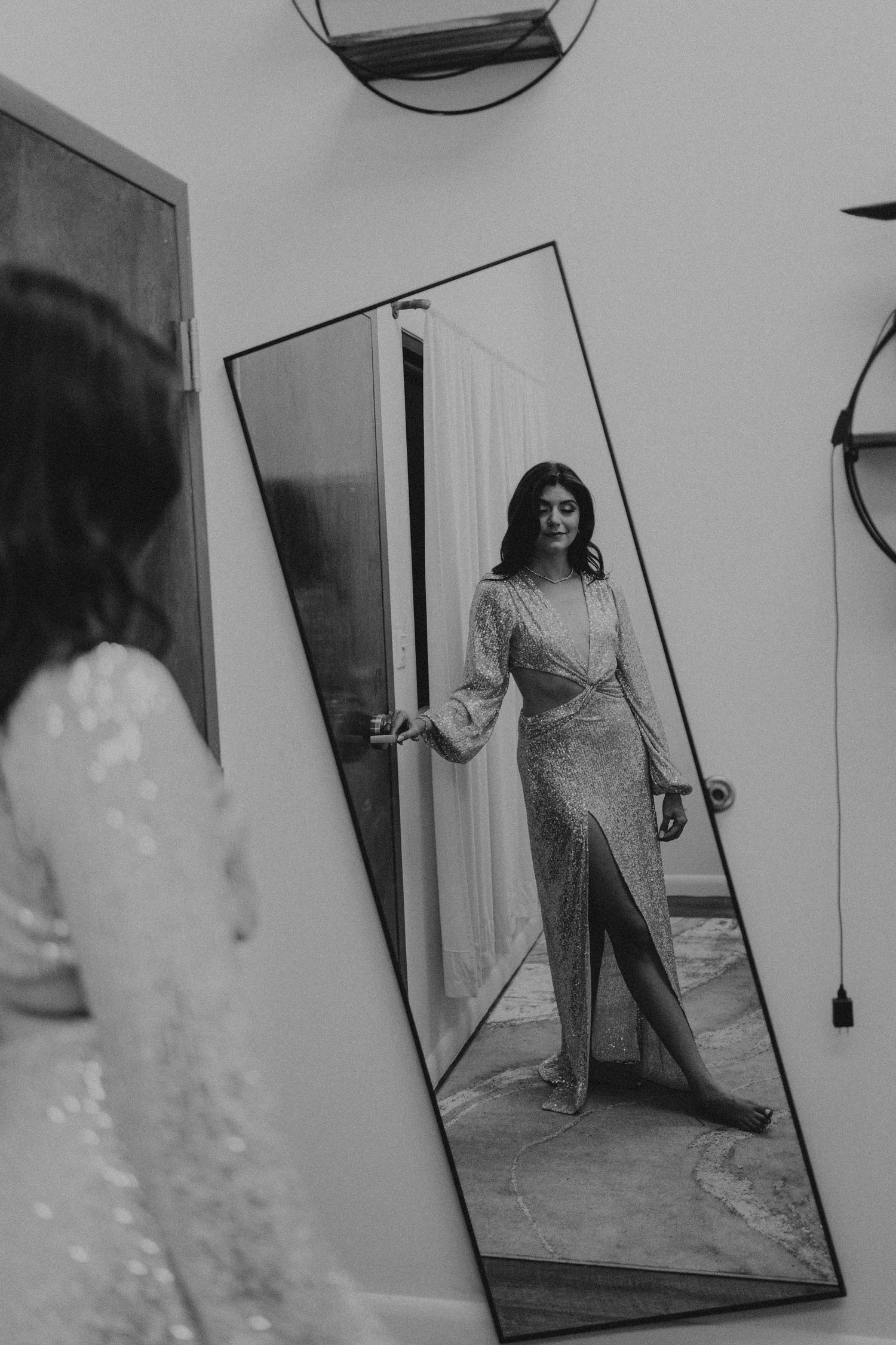 Black and white photo of a bride in a sequined gown with a slit, admiring her reflection in a tall mirror.
