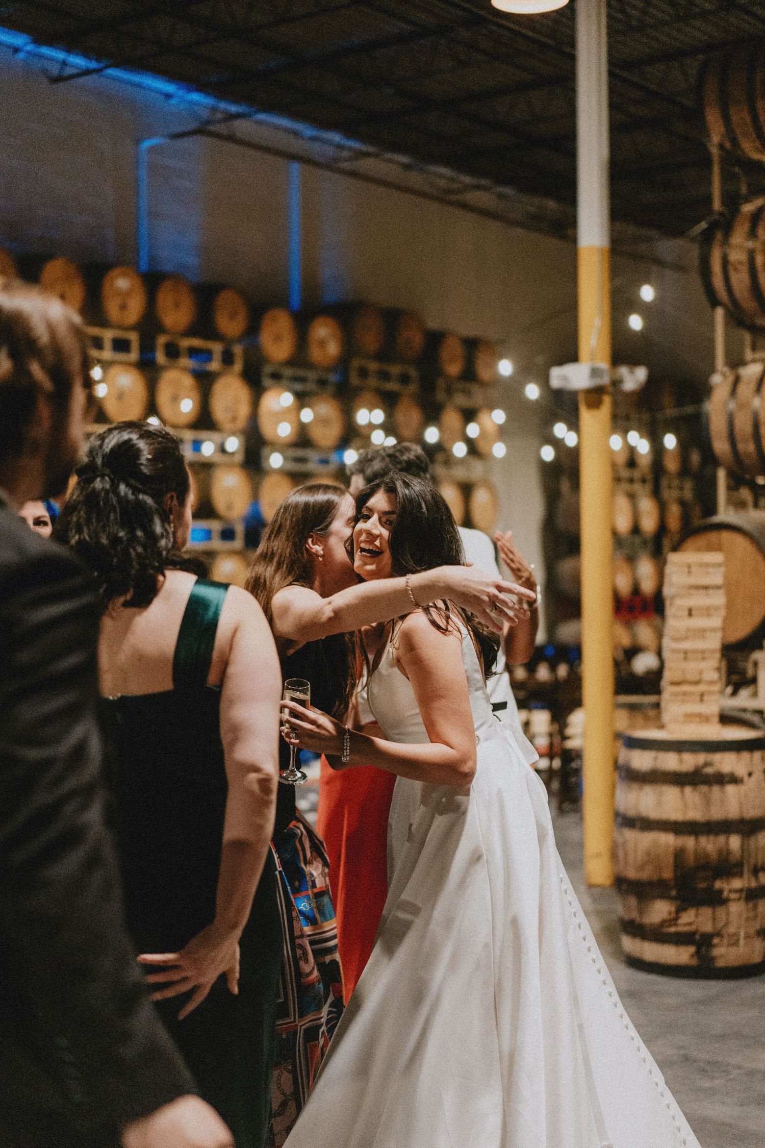 Bride in white gown hugging guests at a rustic wedding reception venue with barrel walls and string lights.
