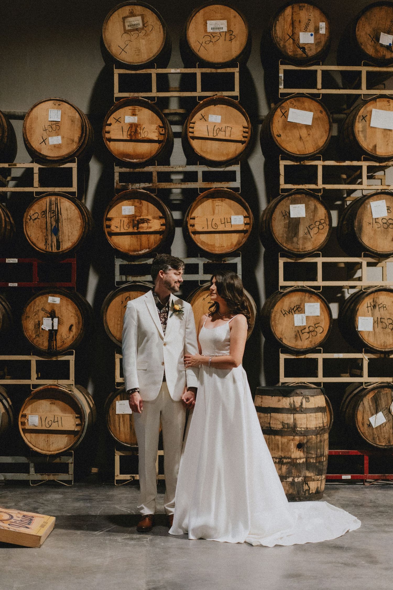 Bride and groom pose romantically in front of stacked wooden whiskey barrels at a distillery wedding venue.