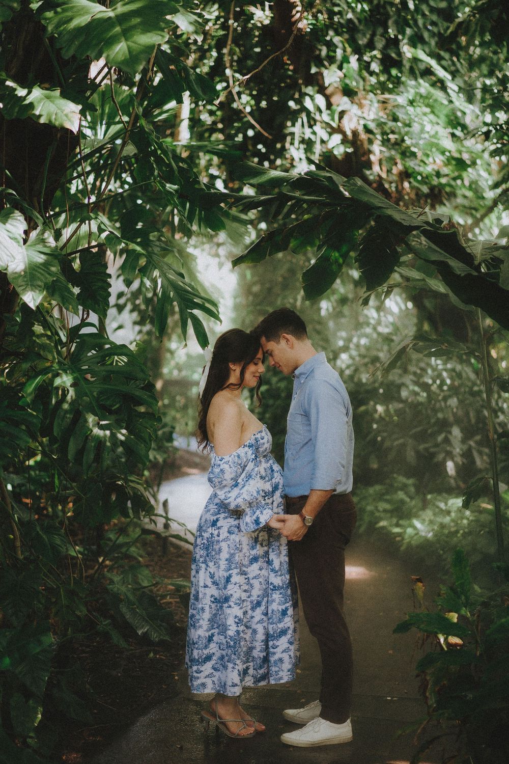 A couple shares an intimate moment on a garden path surrounded by lush greenery and dappled sunlight.