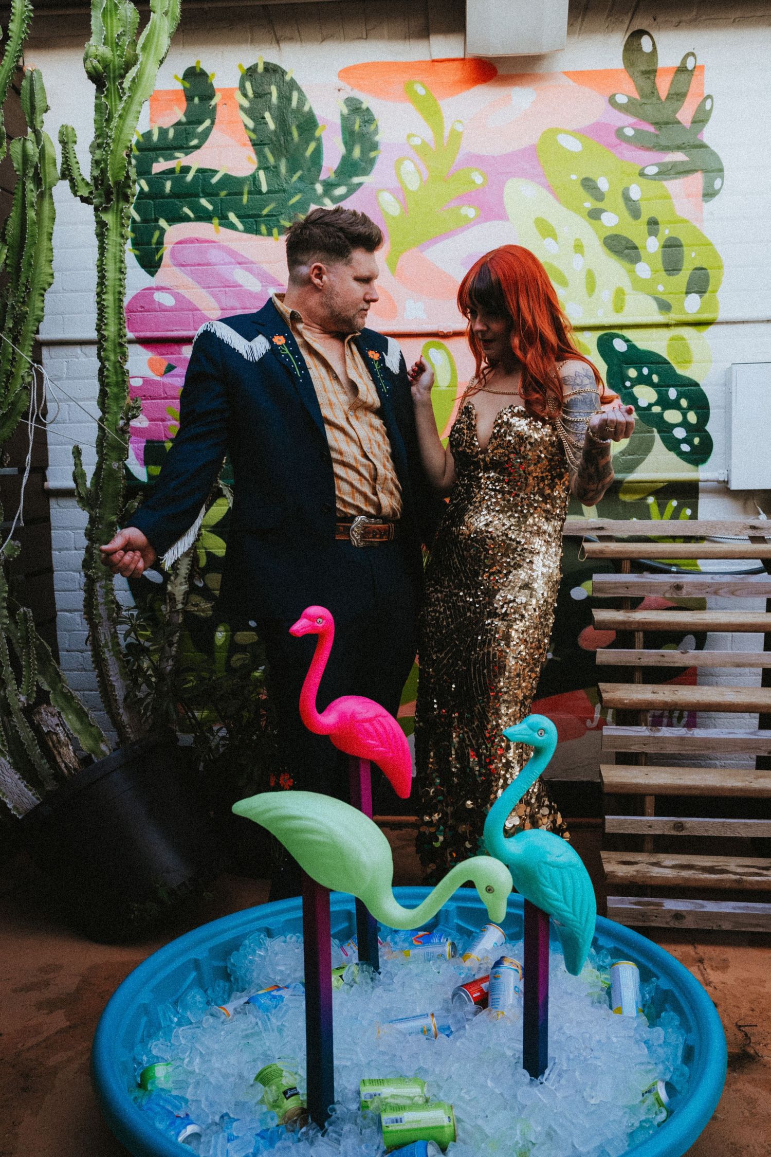 Couple in gold sequin and navy attire posing by a kiddie pool with flamingo decorations and tropical mural.