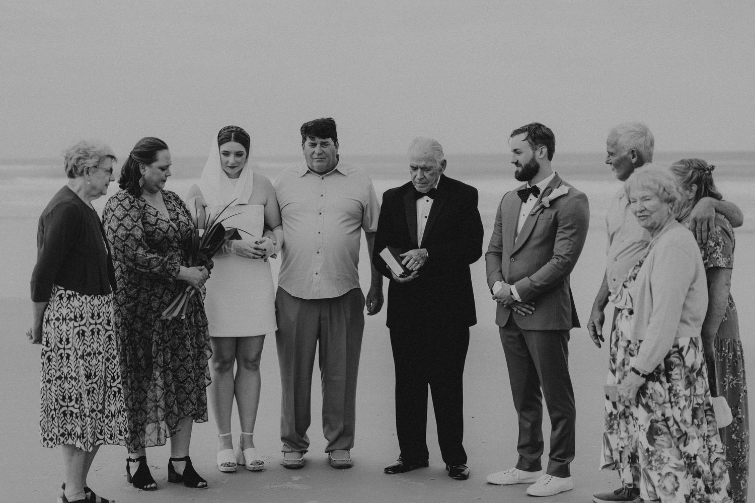 Black and white photo of a wedding group gathered outdoors on a beach, posing together during a ceremony.