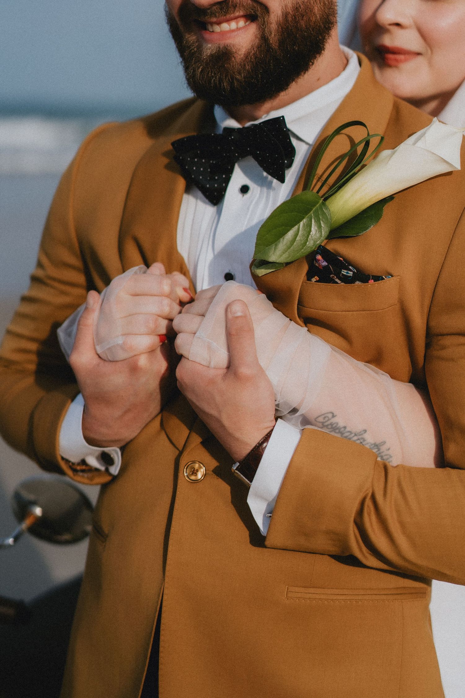 Groom in mustard suit with boutonniere holds brides hands at beach wedding ceremony.