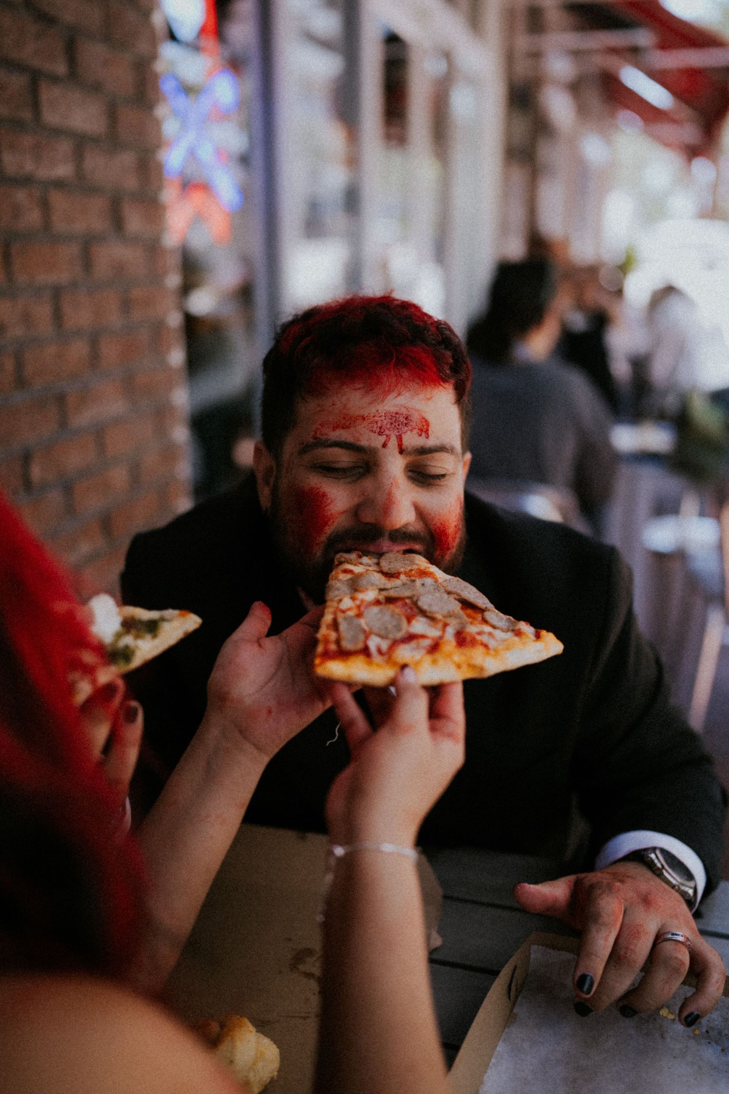 A man in a suit with red face paint has pizza fed to him by a hand at an outdoor restaurant table.