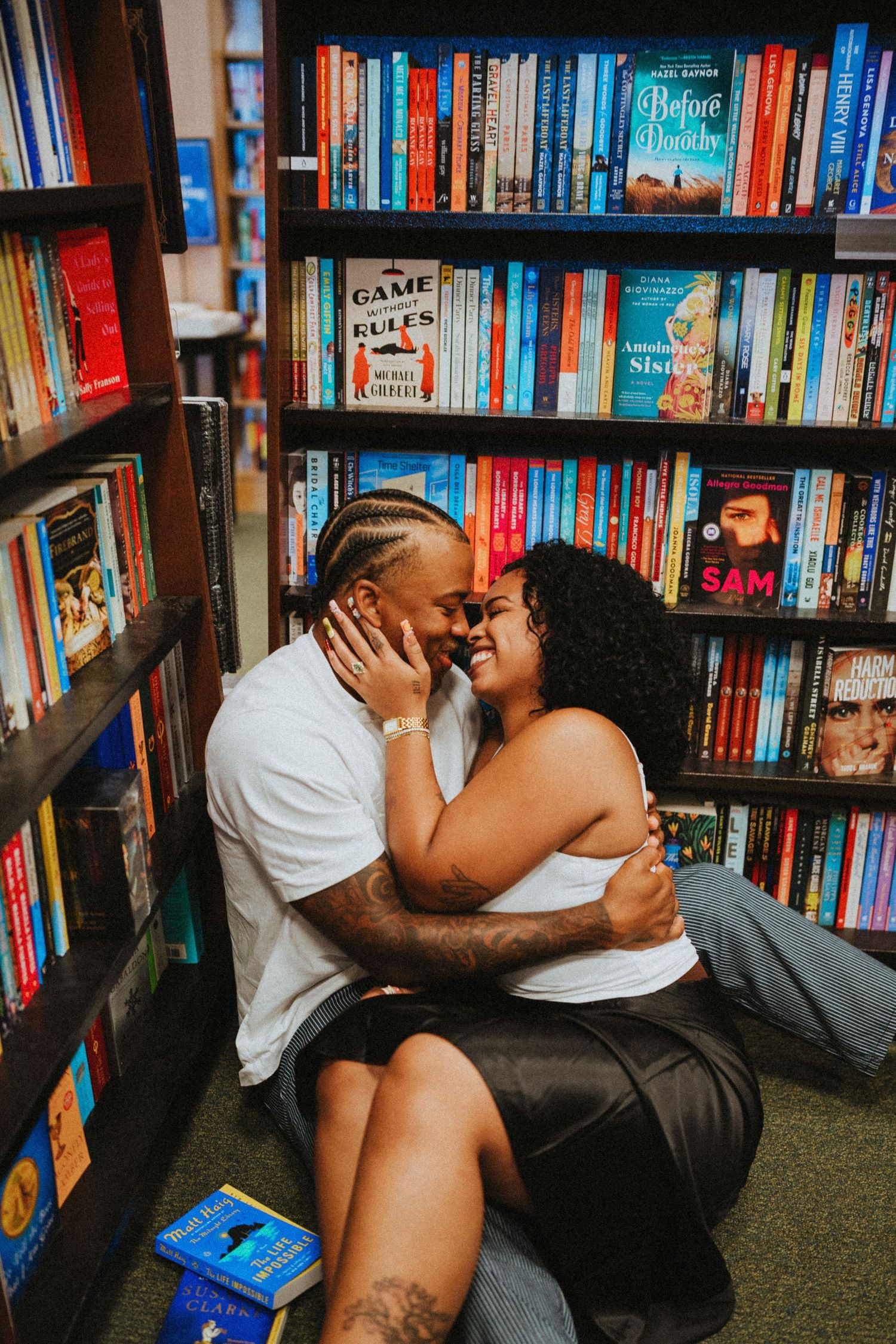 Couple sitting on floor embracing and smiling among colorful bookshelves in a cozy bookstore.