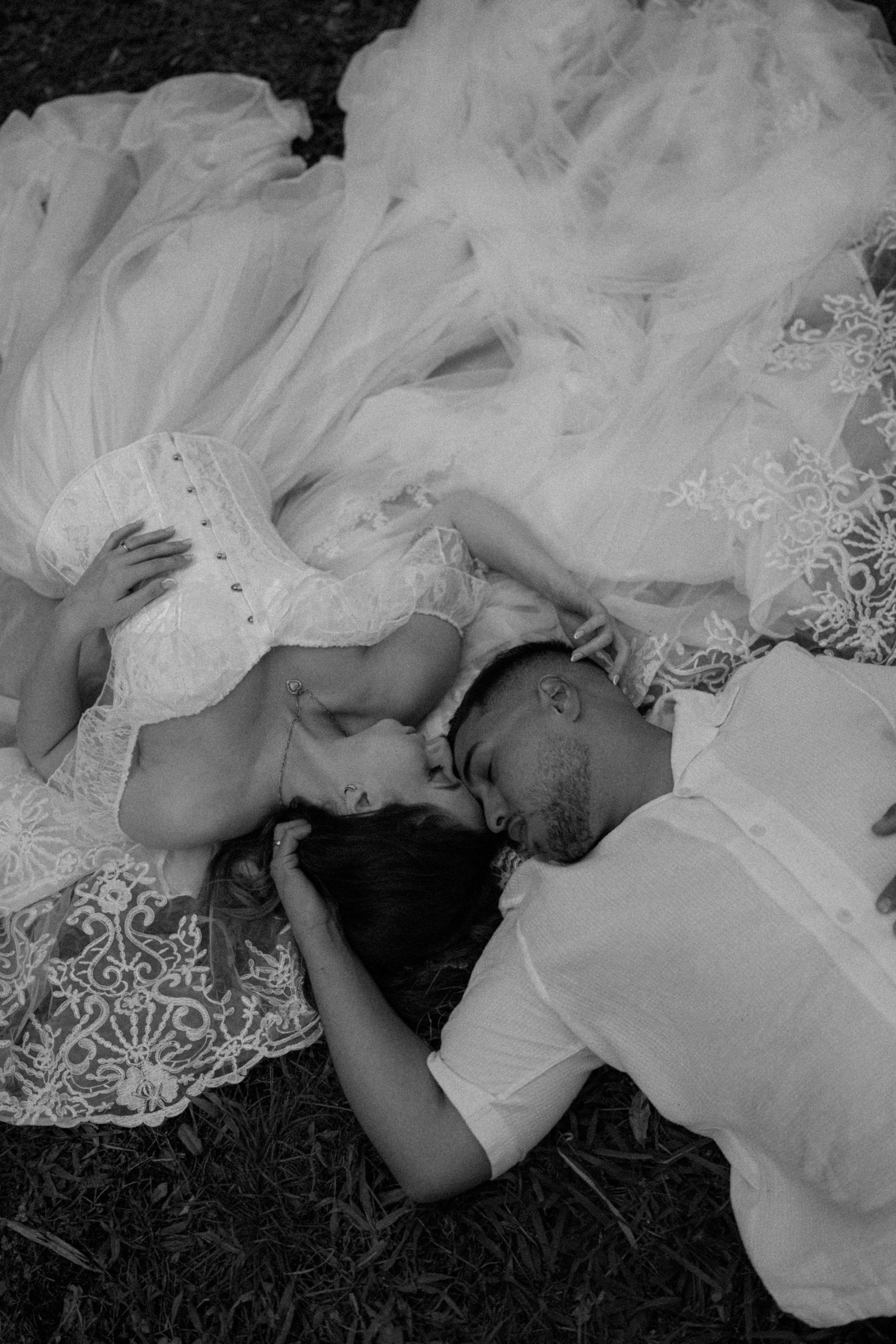 Black and white photo of a bride and groom lying on the ground, sharing a romantic kiss on their wedding day.