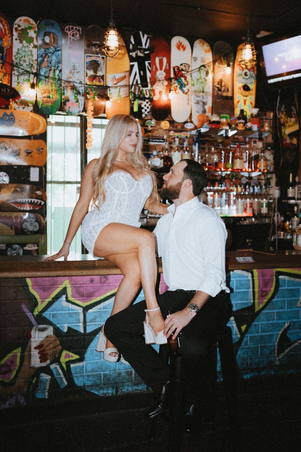 Couple in formal attire poses intimately at bar decorated with skateboard art and graffiti.