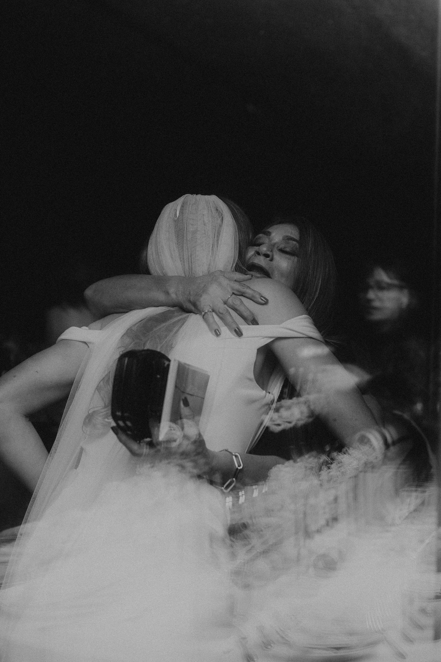 Bride and guest share emotional hug on dance floor surrounded by fog at wedding reception.