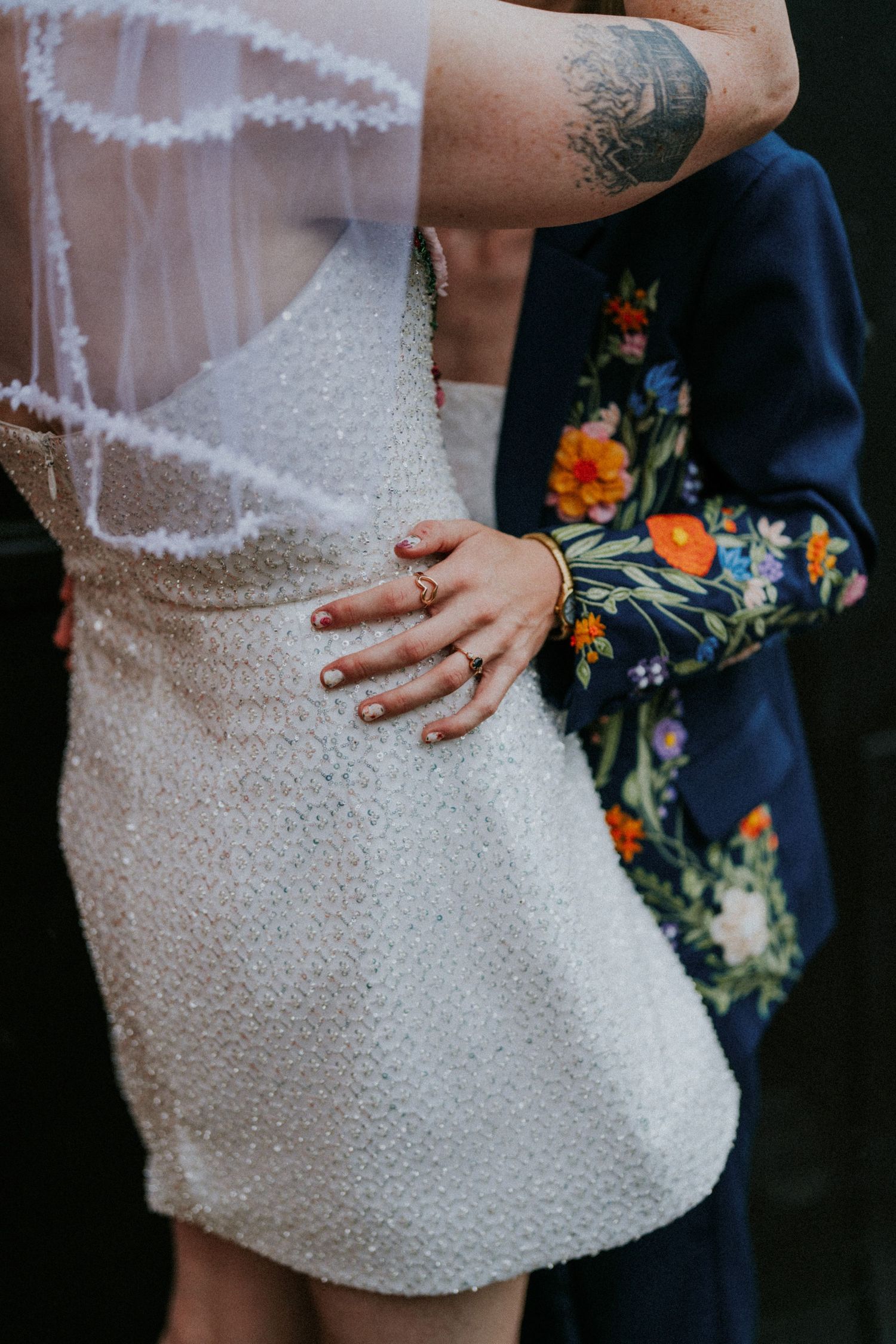 Close-up of bride in white beaded mini dress with groom in floral embroidered navy blazer, wedding rings visible.