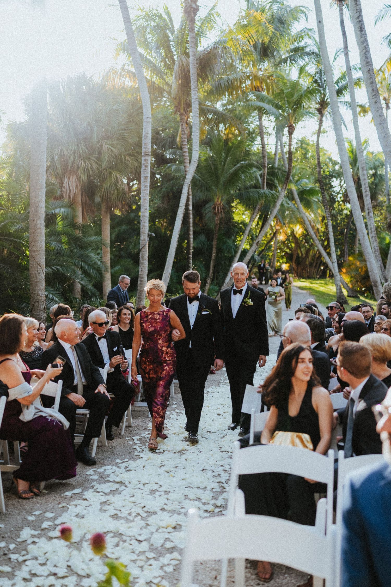 Groom walks down outdoor wedding aisle with parents amid palm trees, white chairs, and rose petal-lined path.