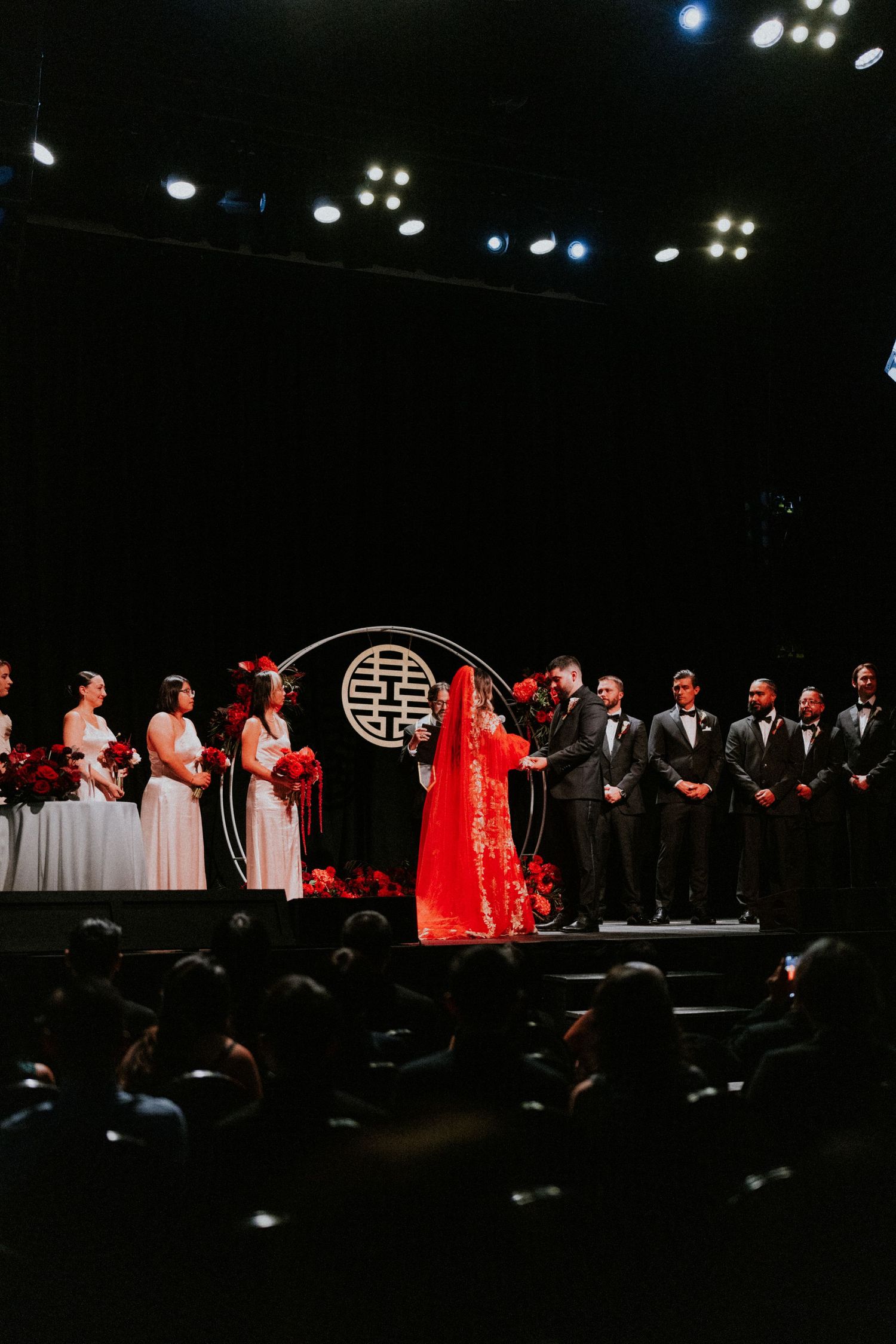 Chinese wedding ceremony on stage with bride in red gown, bridesmaids, groomsmen, and decorative double happiness symbol.