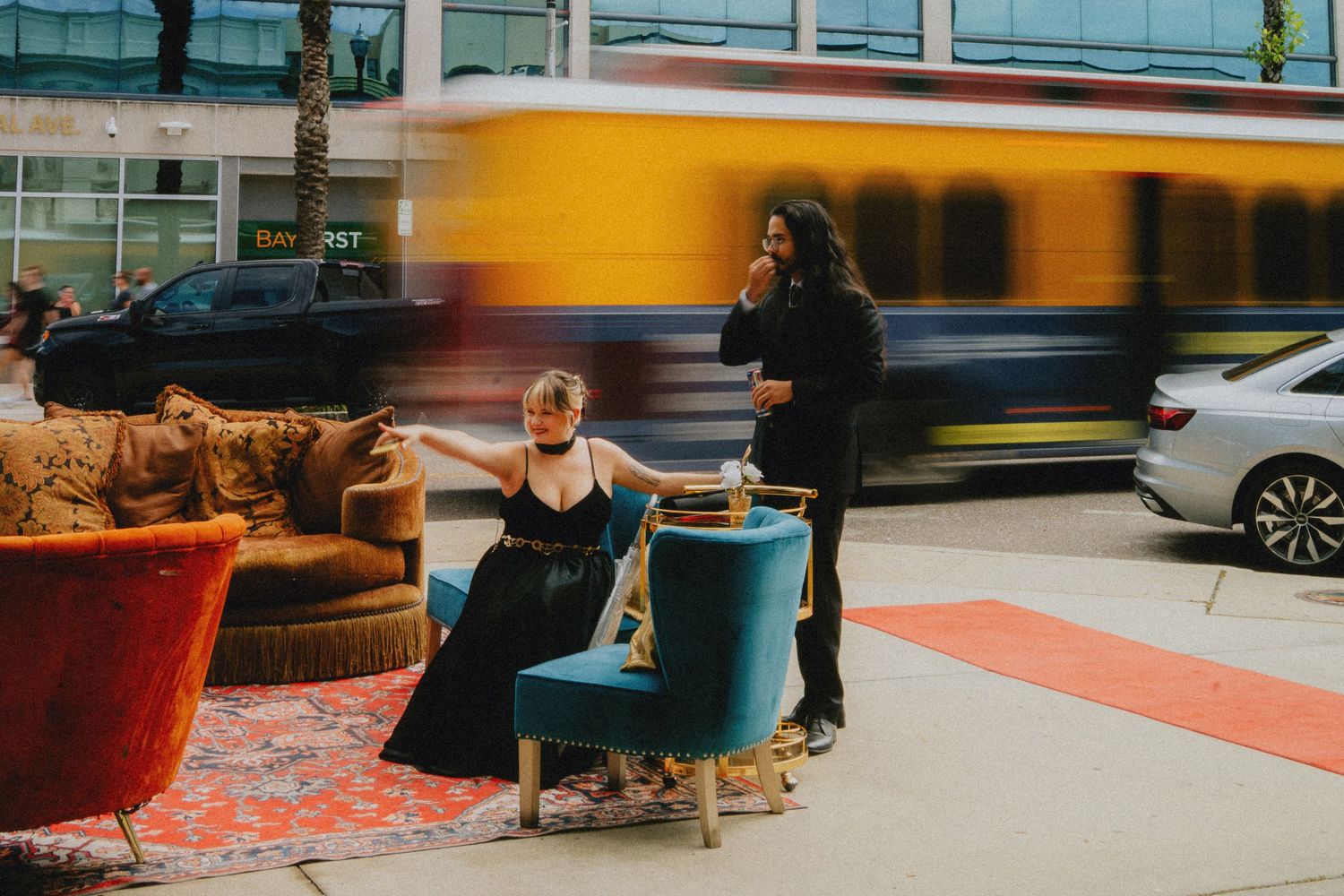 Two people in formal attire sit and stand outdoors among colorful vintage furniture as a yellow bus speeds past.