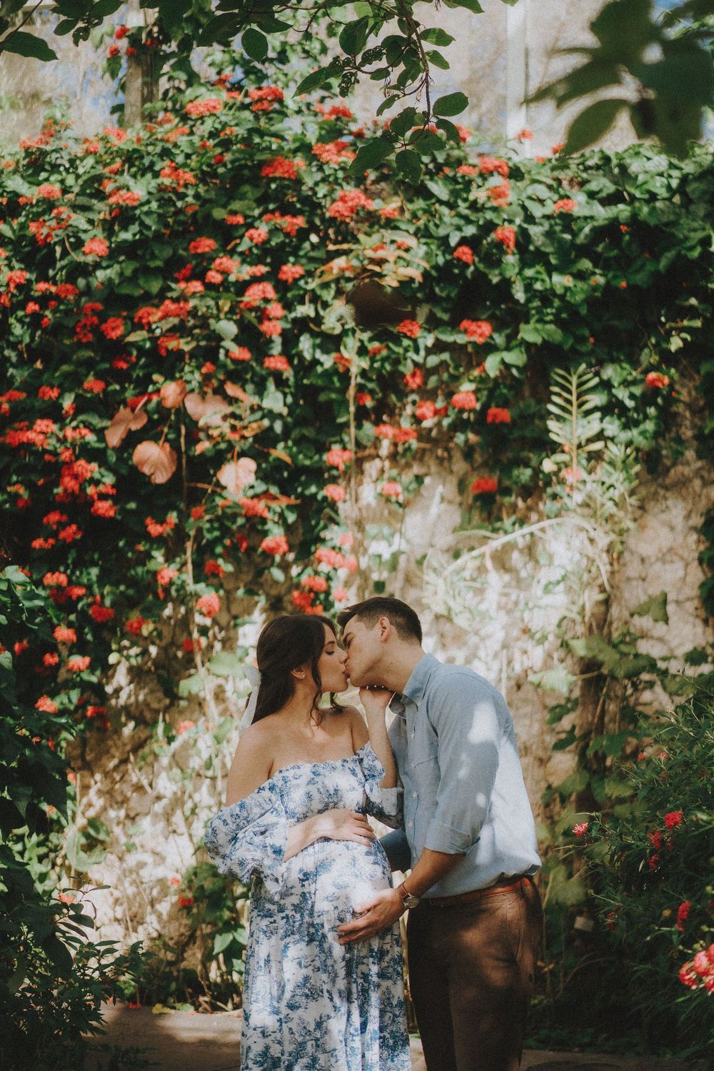 A romantic couple shares tender moments against a backdrop of vibrant red flowers and stone wall in a garden setting.