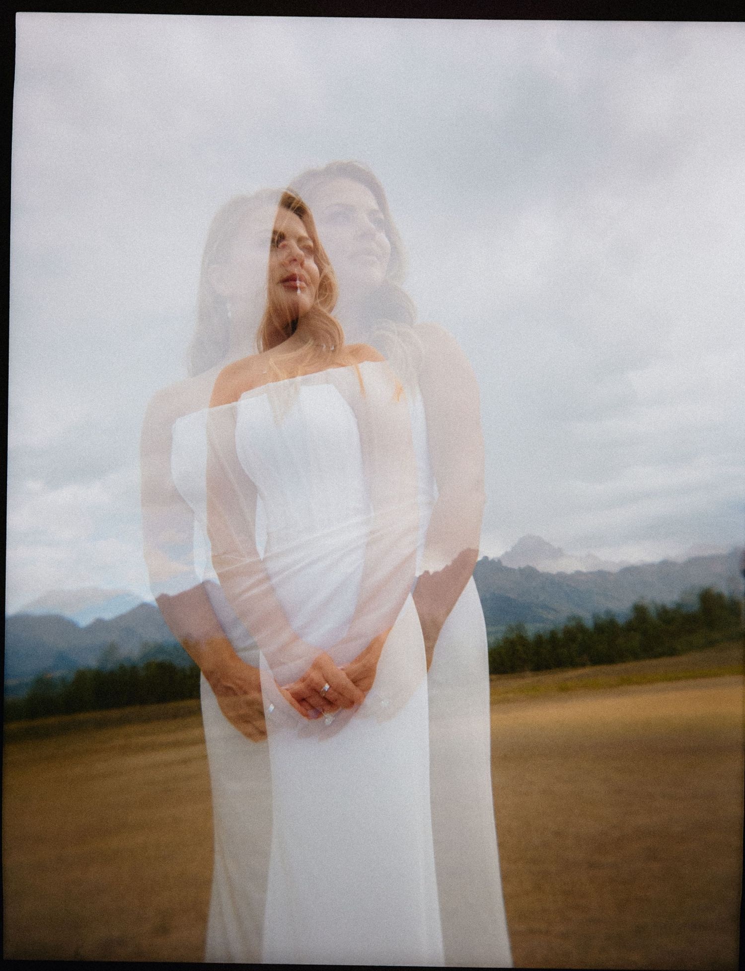 Double exposure photo of a bride in a white gown and veil, standing in a mountain meadow under cloudy skies.