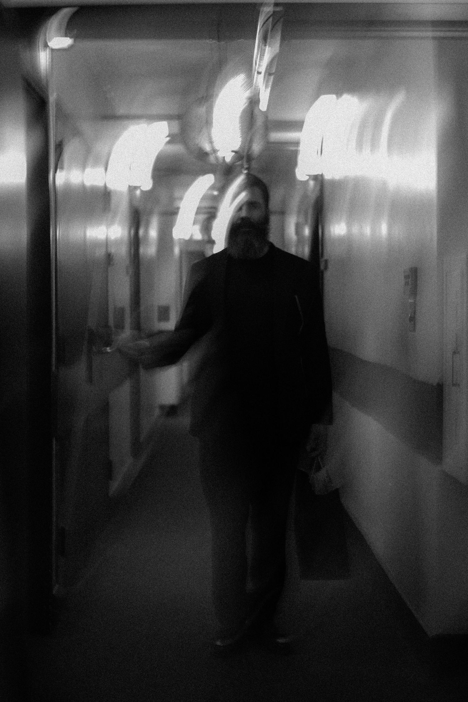 Blurry black and white photo of a bearded man standing in a dimly lit hallway with glowing lights.