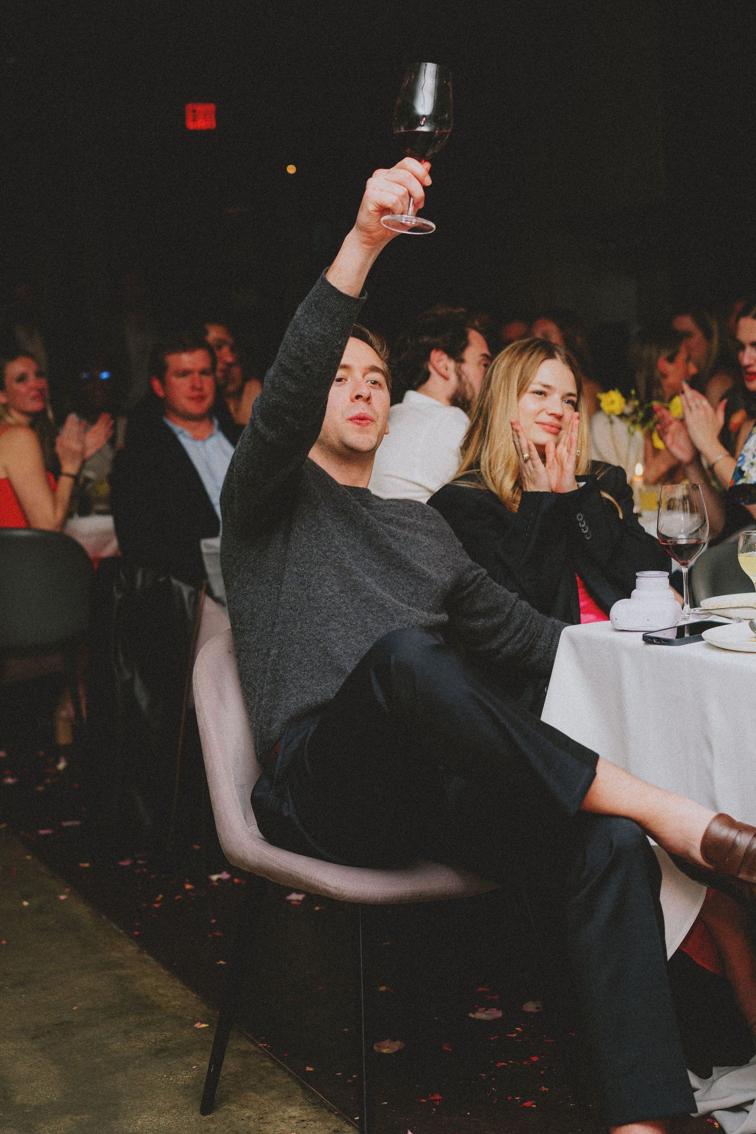Man raising a glass of red wine at an elegant dinner party event with guests seated at white tablecloth tables.
