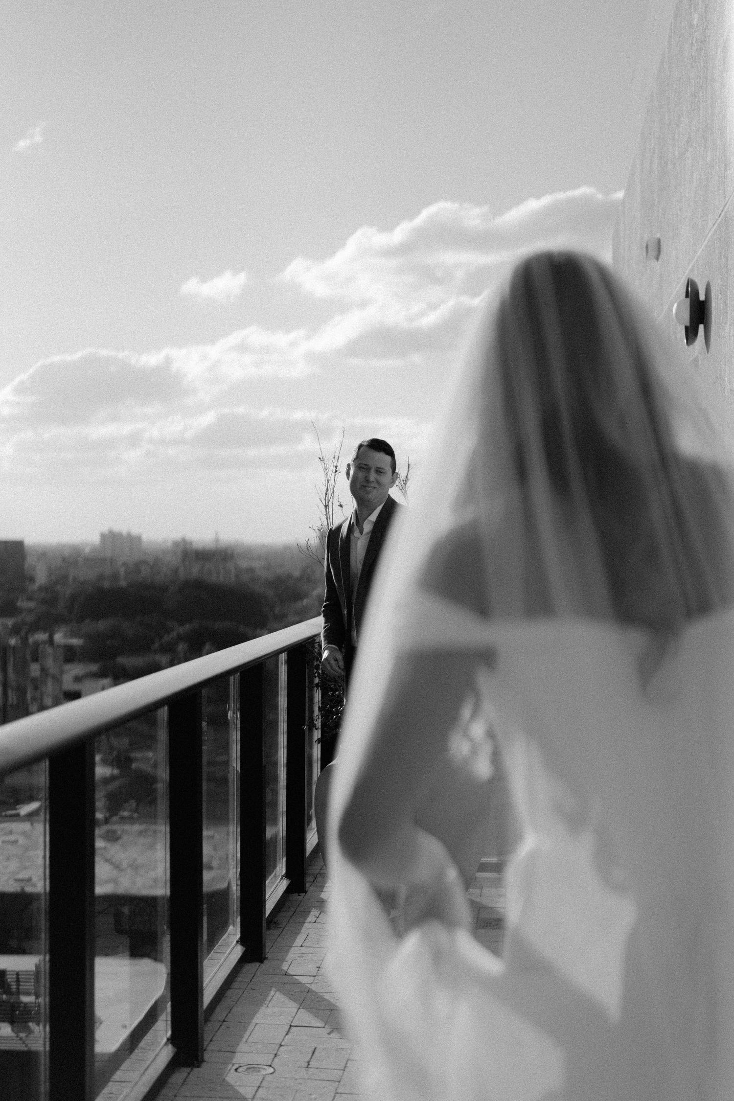 Black and white photo of a groom smiling at his veiled bride on a rooftop balcony with city views.
