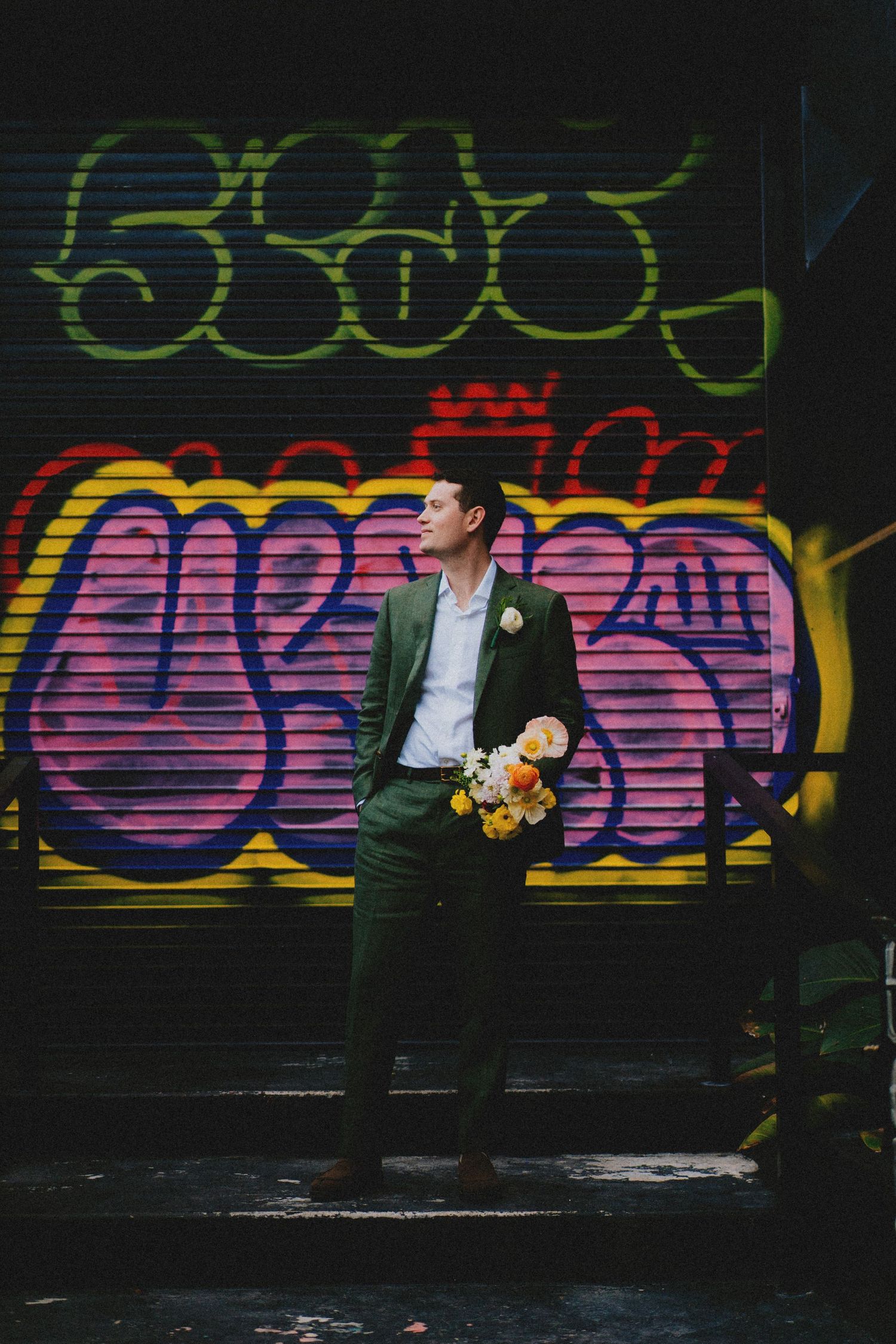 A groom in a green suit holds a colorful bouquet while standing in front of vibrant graffiti wall art.