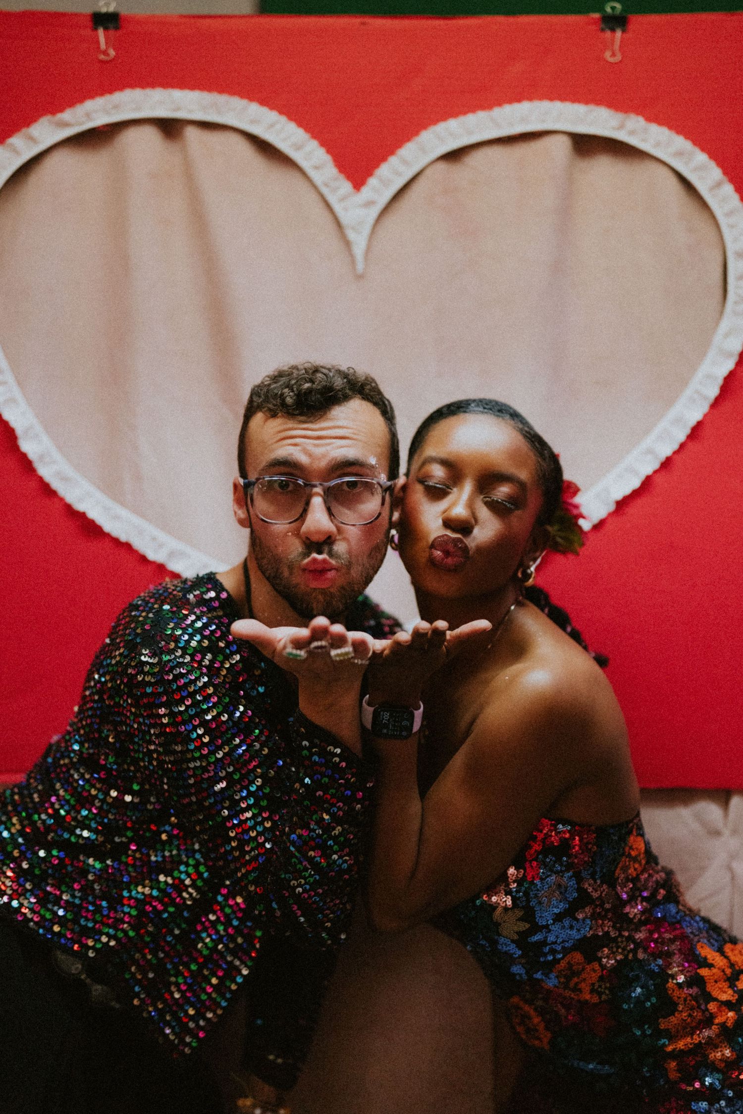 Couple in sequin outfits blowing kisses in front of a red heart backdrop at a festive event.