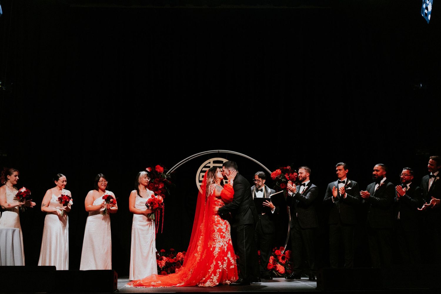Bride in red gown with wedding party on stage with circular arch and red bouquets.