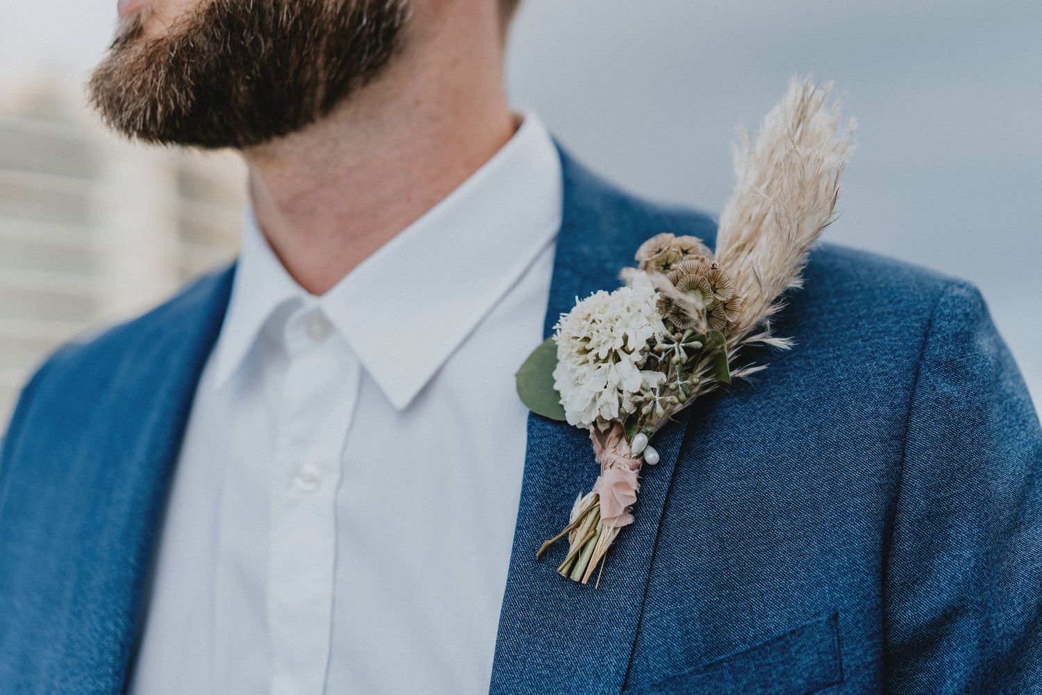 Close-up of a groom in a blue suit wearing a boho dried flower boutonniere with pampas grass and pink blooms.