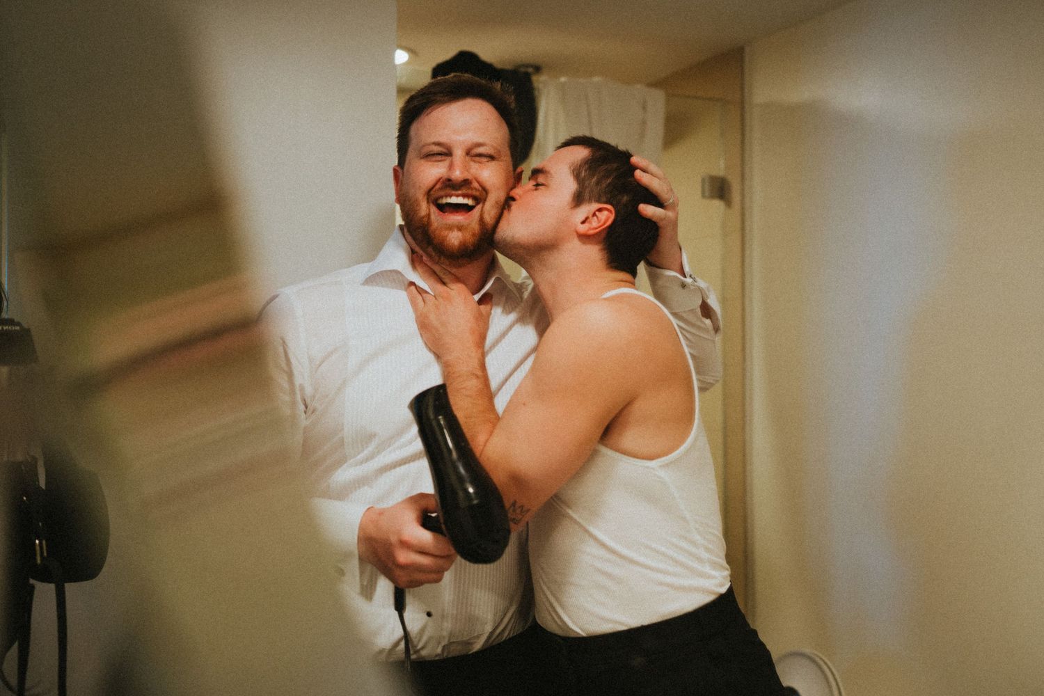 Two men laughing and sharing a joyful moment in a bathroom, one holding a hair dryer while the other kisses his cheek.