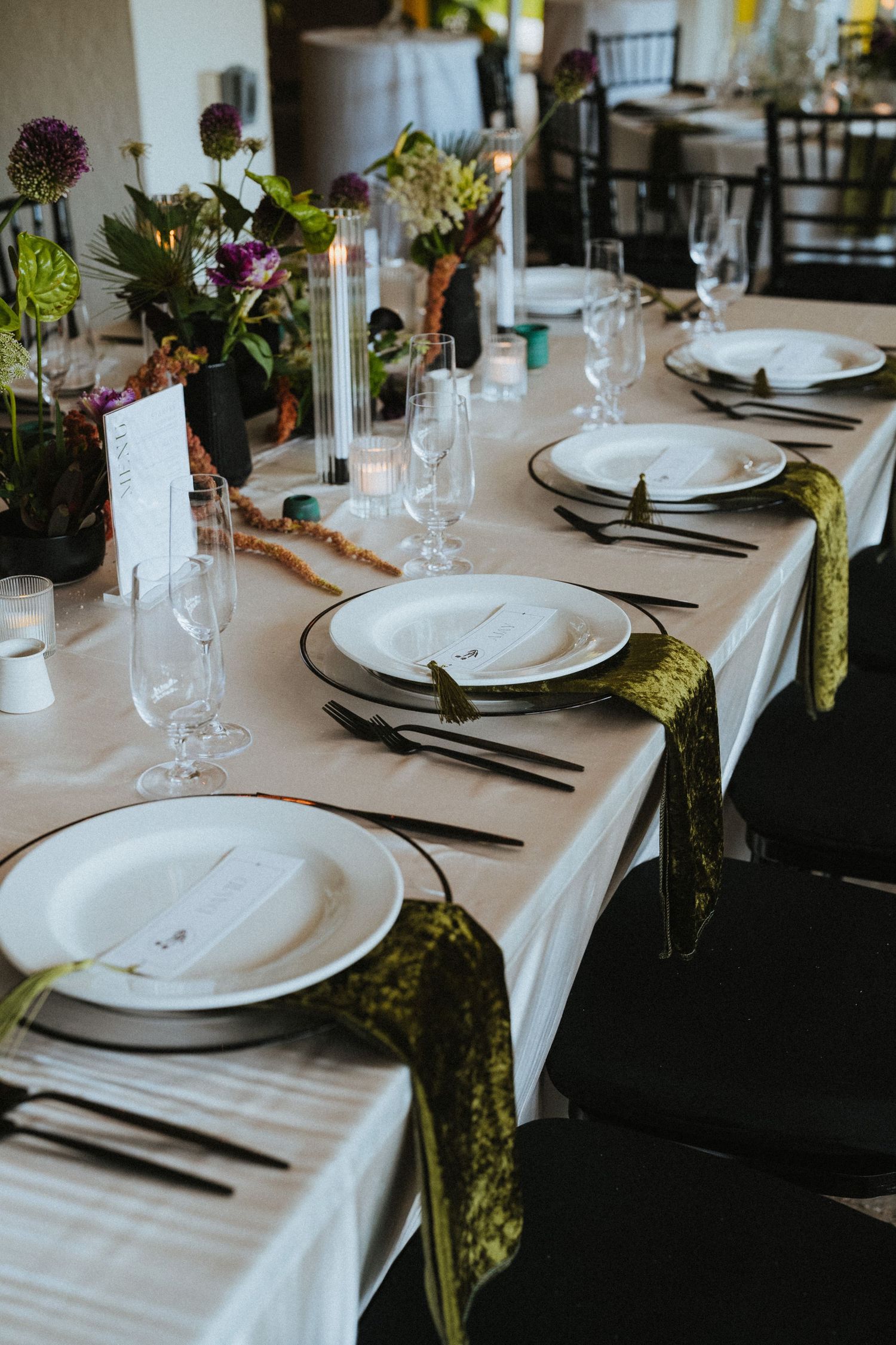 Elegant wedding reception table set with white plates, green velvet napkins, black cutlery, and floral centerpieces.