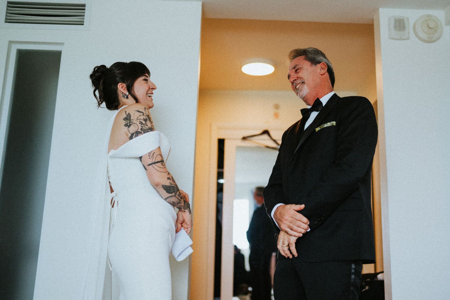 Bride in white off-shoulder gown with tattoos shares a joyful first look moment with her father in a tuxedo.