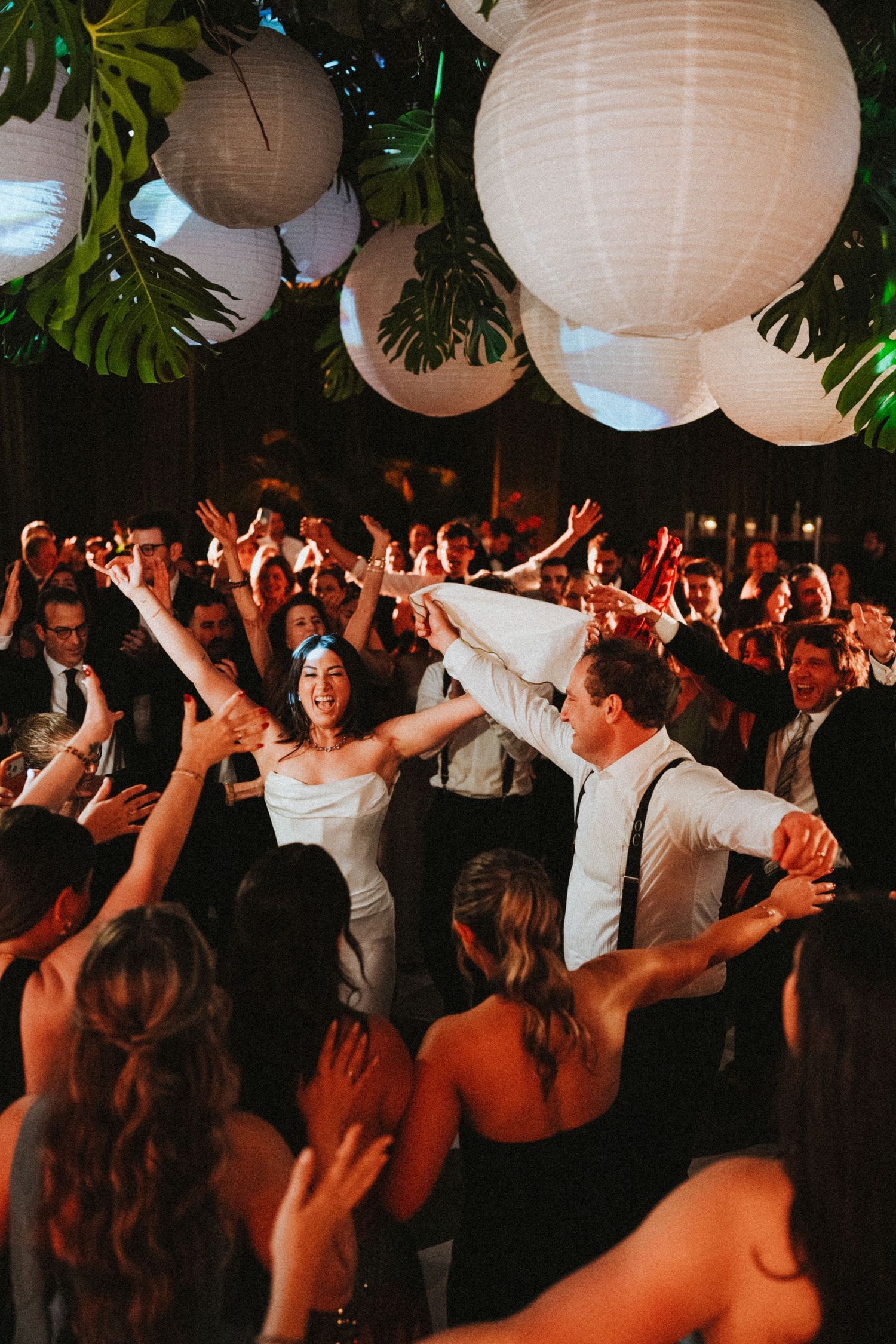 Bride and groom celebrate joyfully on a crowded wedding dance floor under white paper lantern decorations.